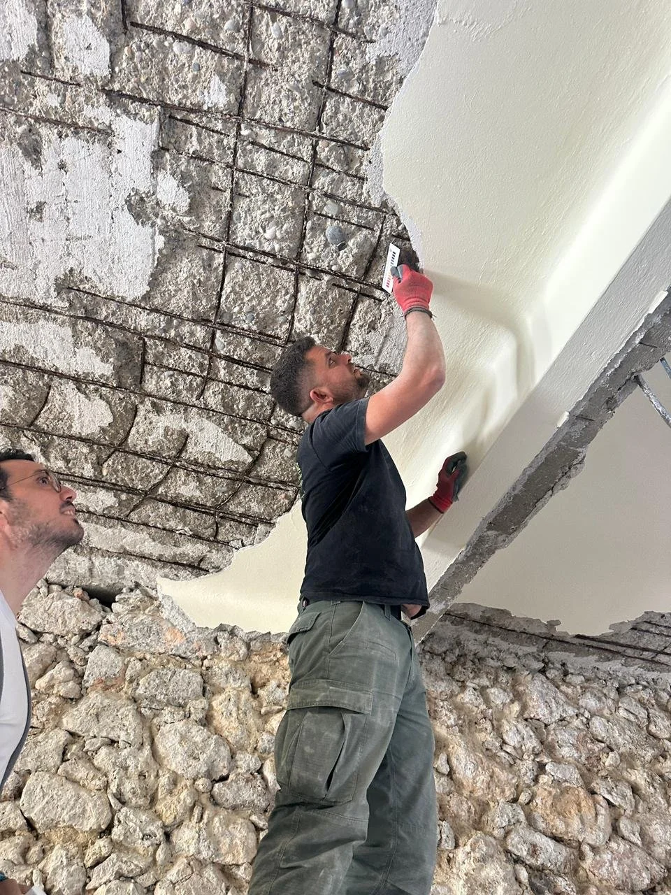Two men working on a construction site with exposed brick and concrete; one man is standing on a pile of rocks and using a tool on the ceiling, while the other man looks on.