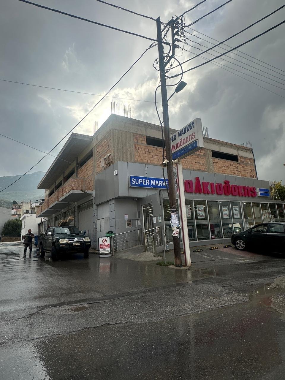 A building with signs reading 'Super Market' and 'ΑΚΡΙΔΟΚΑΣ' on a rainy day. There are two cars parked in front, and a man standing near one of the vehicles, with wet pavement and cloudy sky overhead.