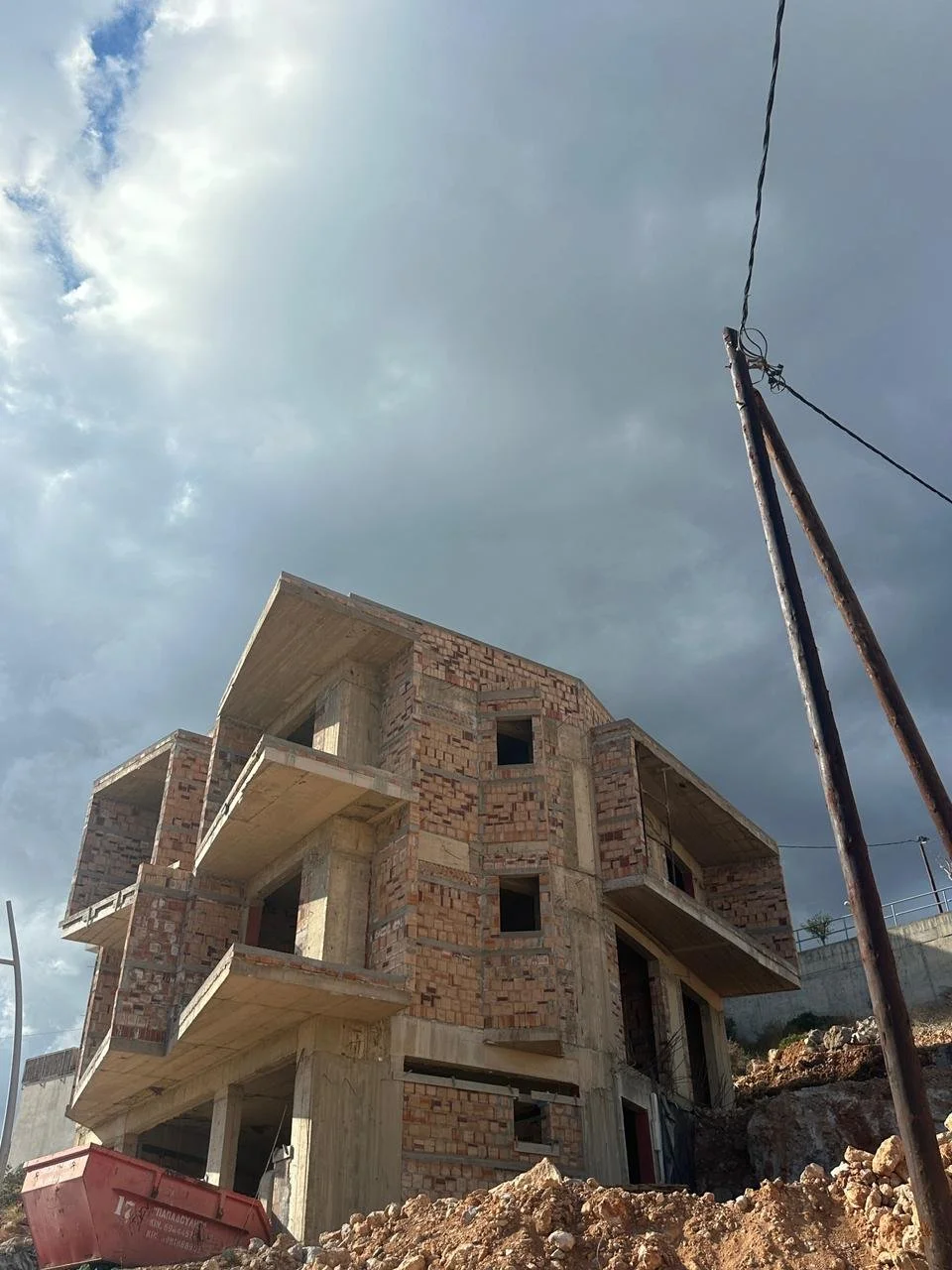 Construction site with an unfinished multi-story brick and concrete building against a cloudy sky, with a damaged utility pole and rubble in the foreground.