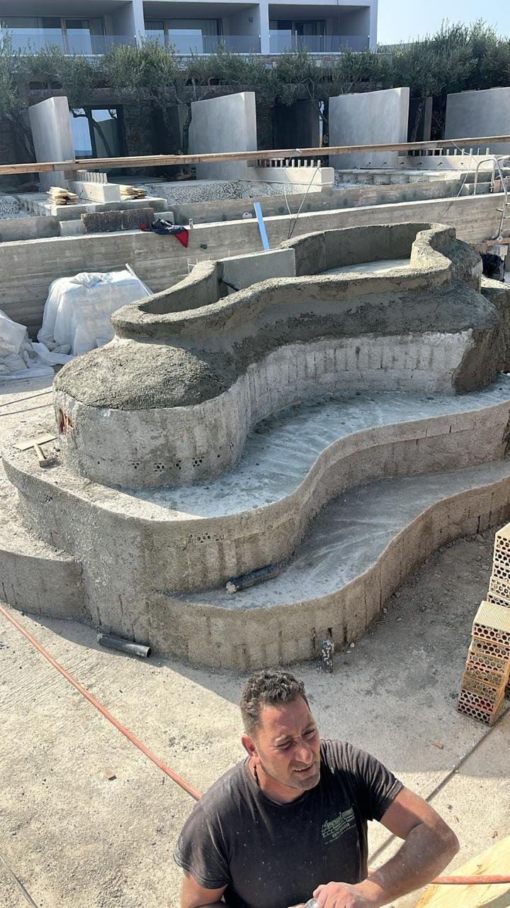 Under construction swimming pool with curved concrete structure, a worker with a black shirt, construction tools, and building materials around.