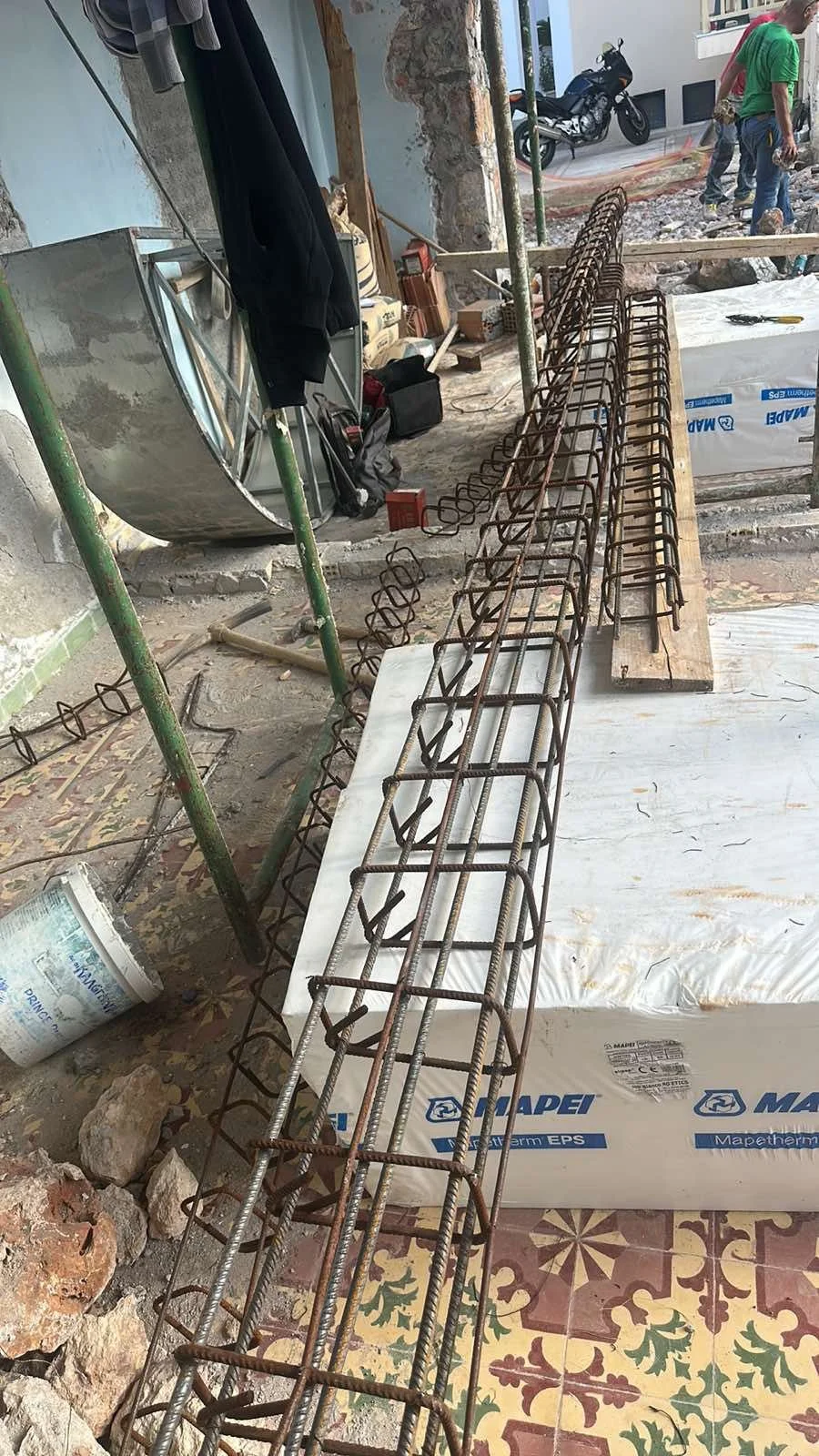 Construction site with exposed steel rebar, tools, and workers in the background. A motorcycle is parked outside.