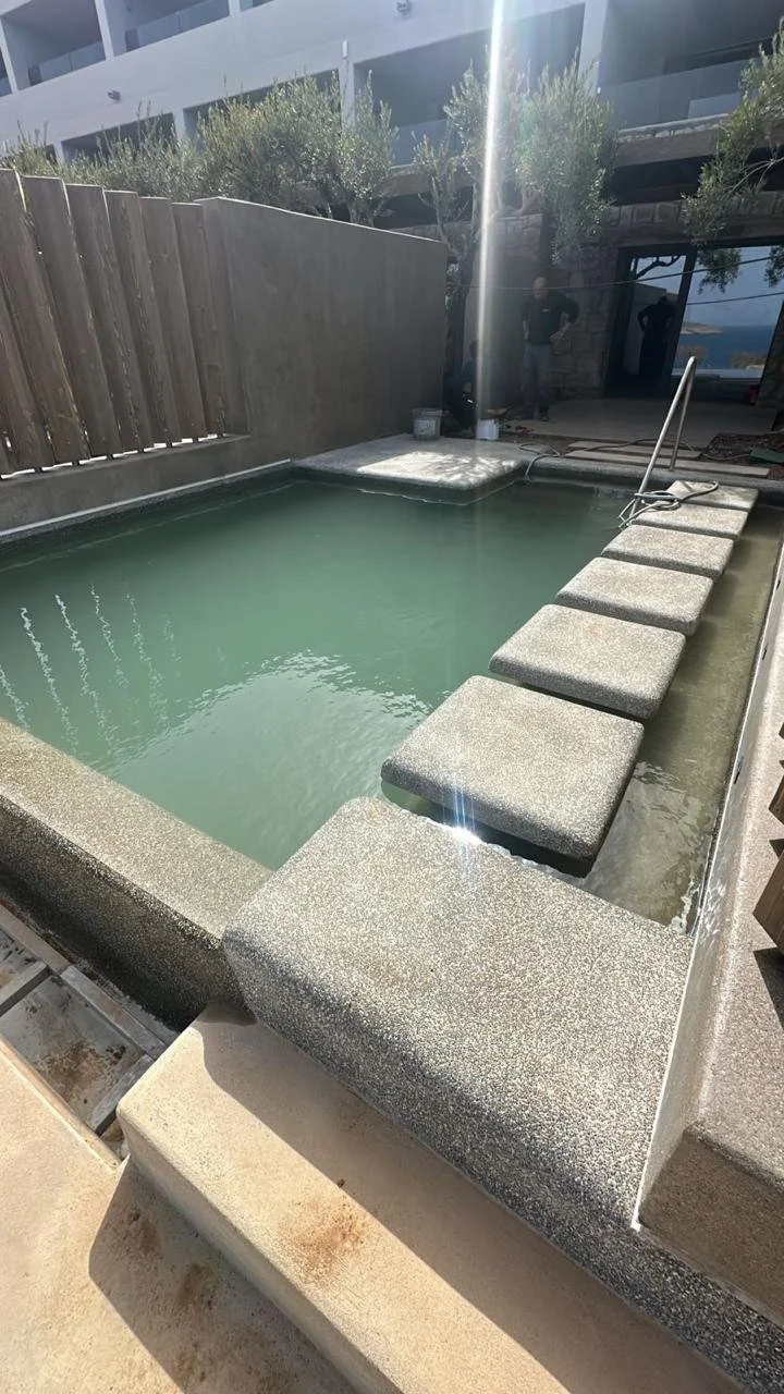 Small rectangular hot tub with concrete seating around the edges, fenced backyard, and a building with trees in the background.