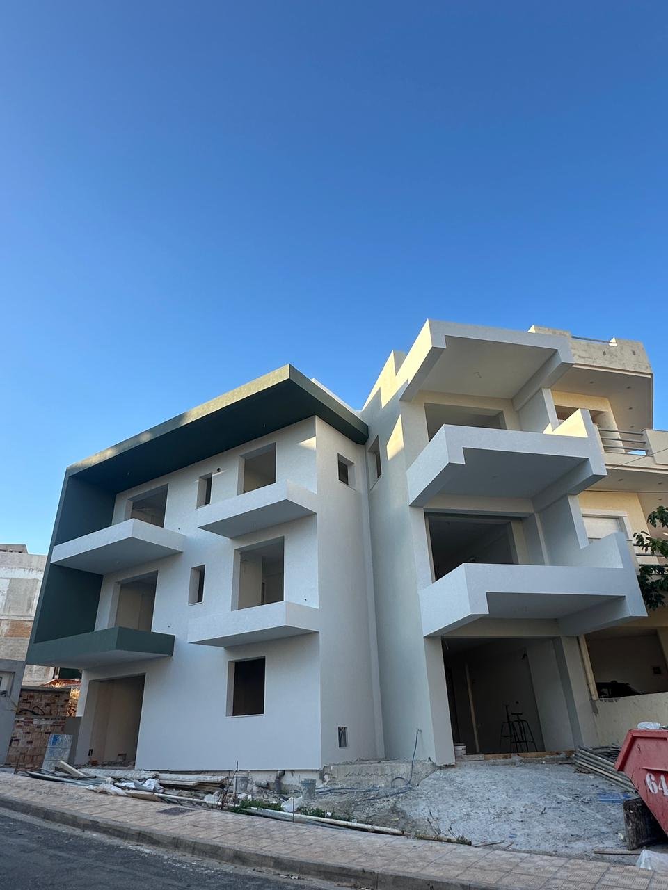 A modern multi-story house under construction with white walls, balconies, and a partly finished exterior, set against a bright blue sky.