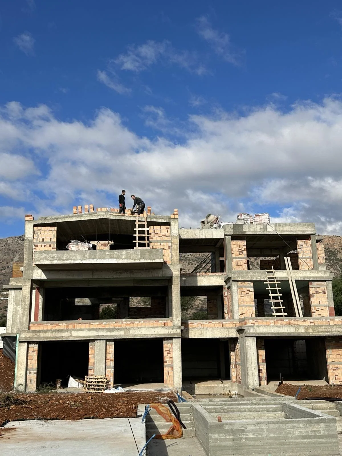 Construction site with a multi-story building under construction, two workers at the top floor, and blue sky with clouds in the background.