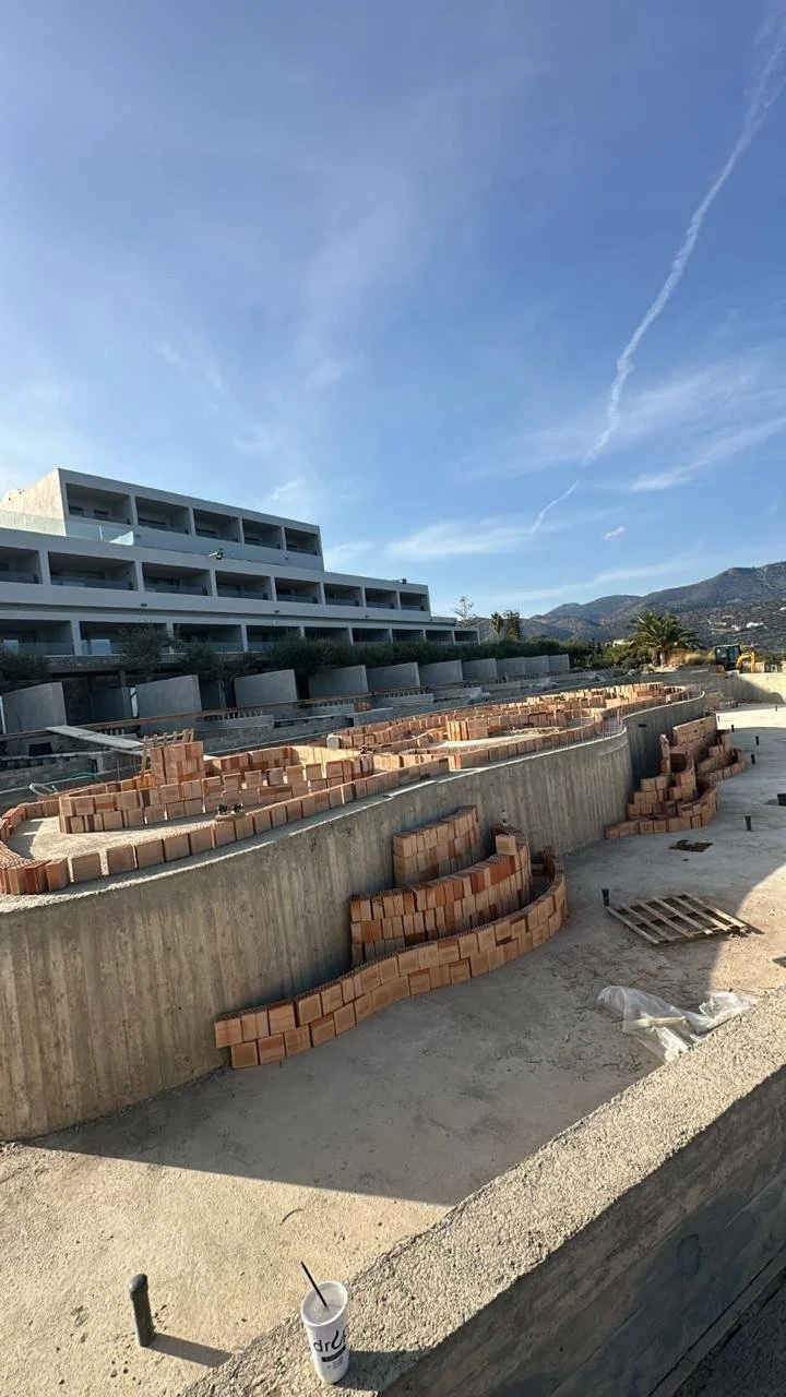 Construction site with brickwork and a modern multi-story building in the background, mountains in the distance, and a clear blue sky.