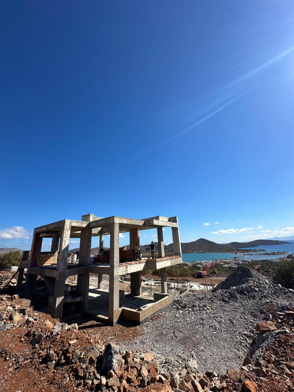 Under construction concrete building frame on a hilltop overlooking a lake and distant mountains, with construction equipment and workers.