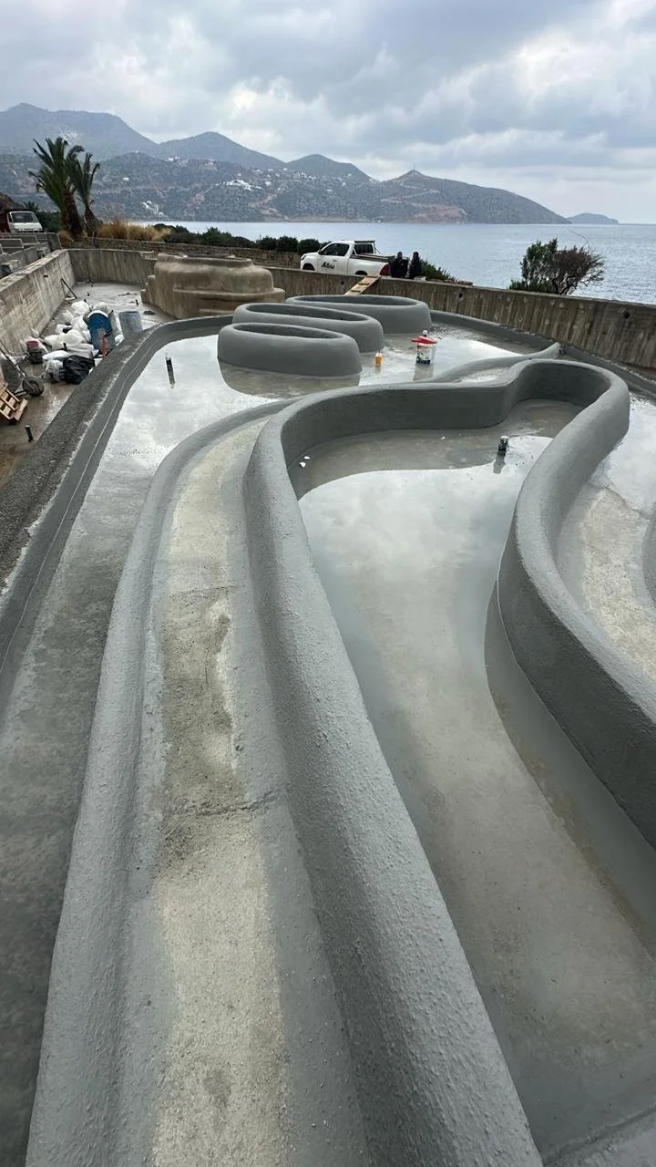 Construction site of a swimming pool overlooking the ocean, with hills in the background, partly cloudy sky, and construction materials nearby.