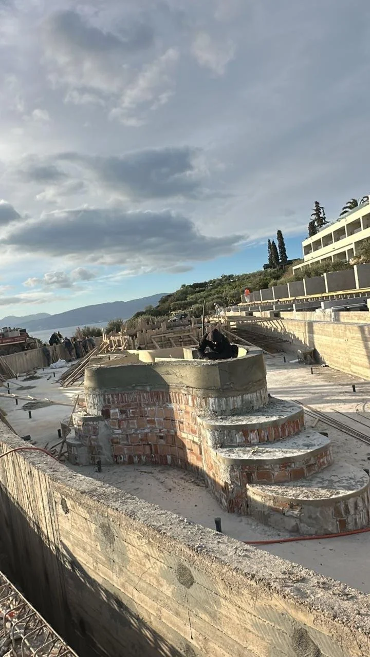 Construction site with a circular brick and concrete structure under construction, set against a backdrop of a cloudy sky and distant mountains, with buildings on a hillside to the right.