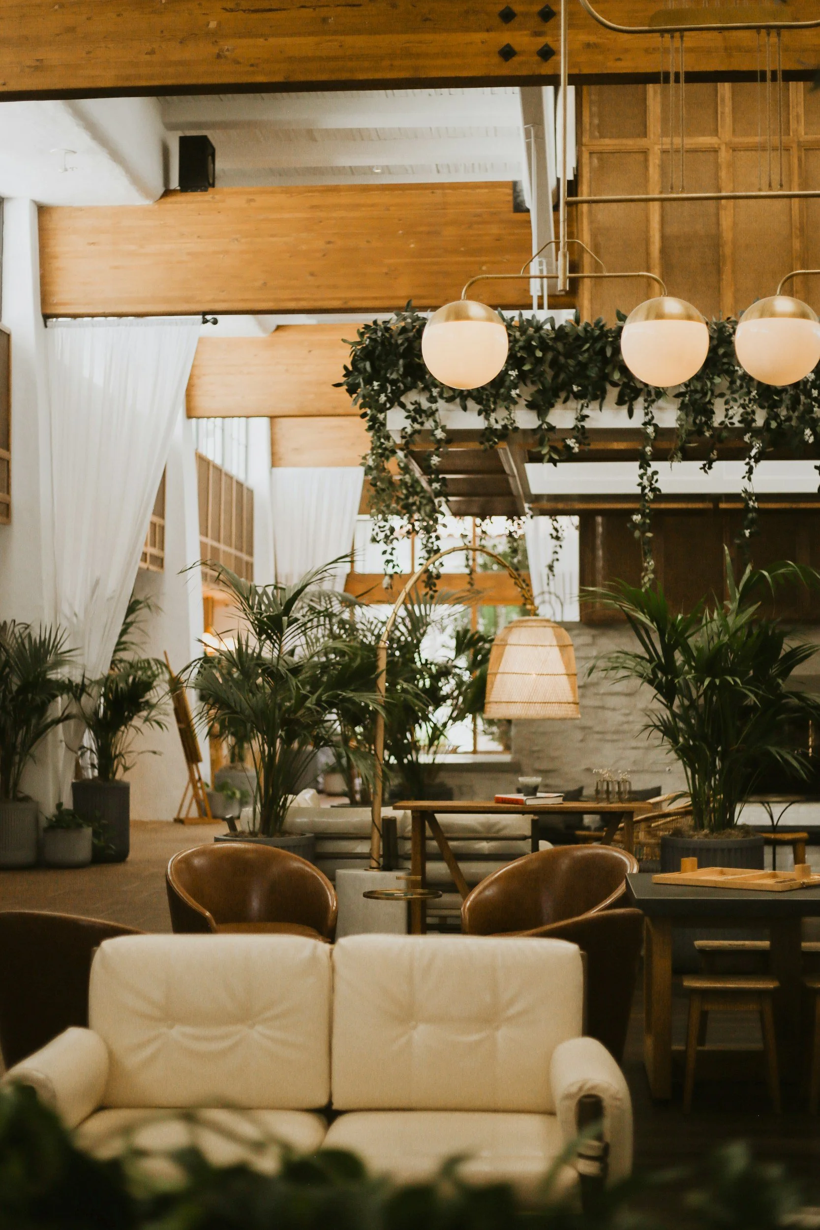 Interior of a cozy, modern living space featuring a cream-colored sofa in the foreground, surrounded by potted plants. In the background, there are wooden chairs, tables, and hanging pendant lights, with lush green plants and decorative elements creating a warm, inviting atmosphere.