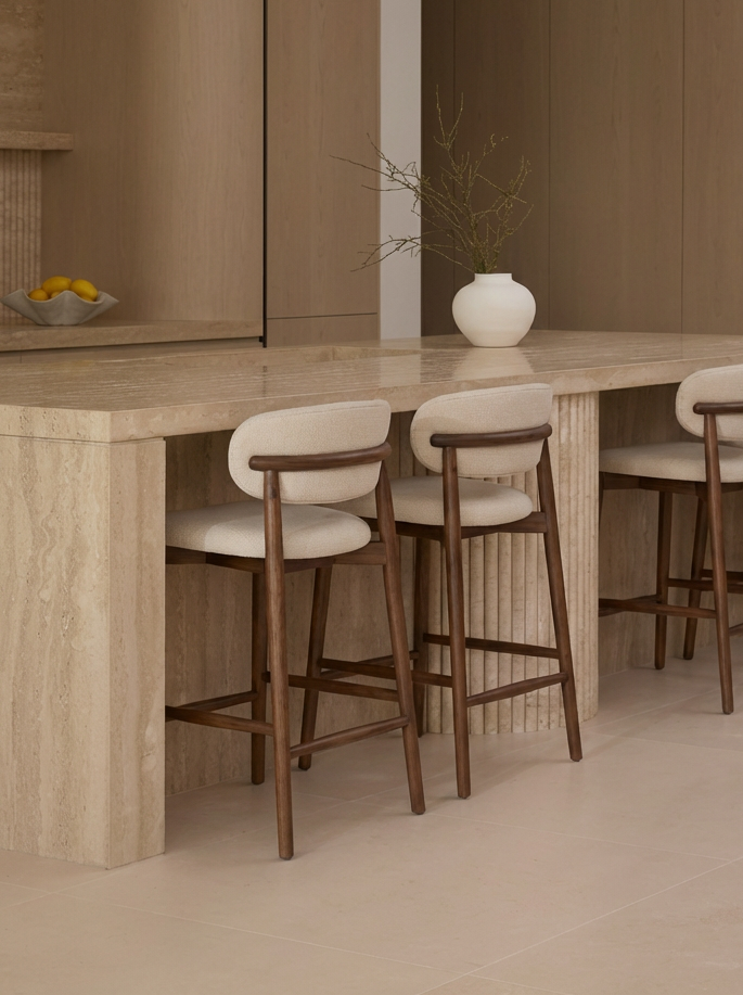 A beige stone kitchen counter with three wooden chairs that have cream upholstered seats, and a white vase with bare branches on the counter.