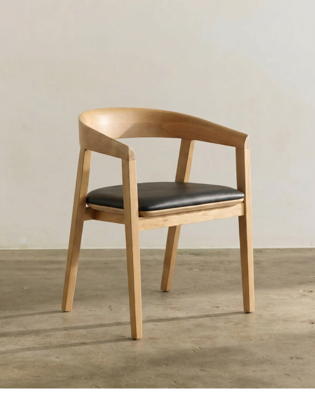 A wooden chair with a curved backrest and black cushioned seat, placed on a concrete floor against a plain wall.
