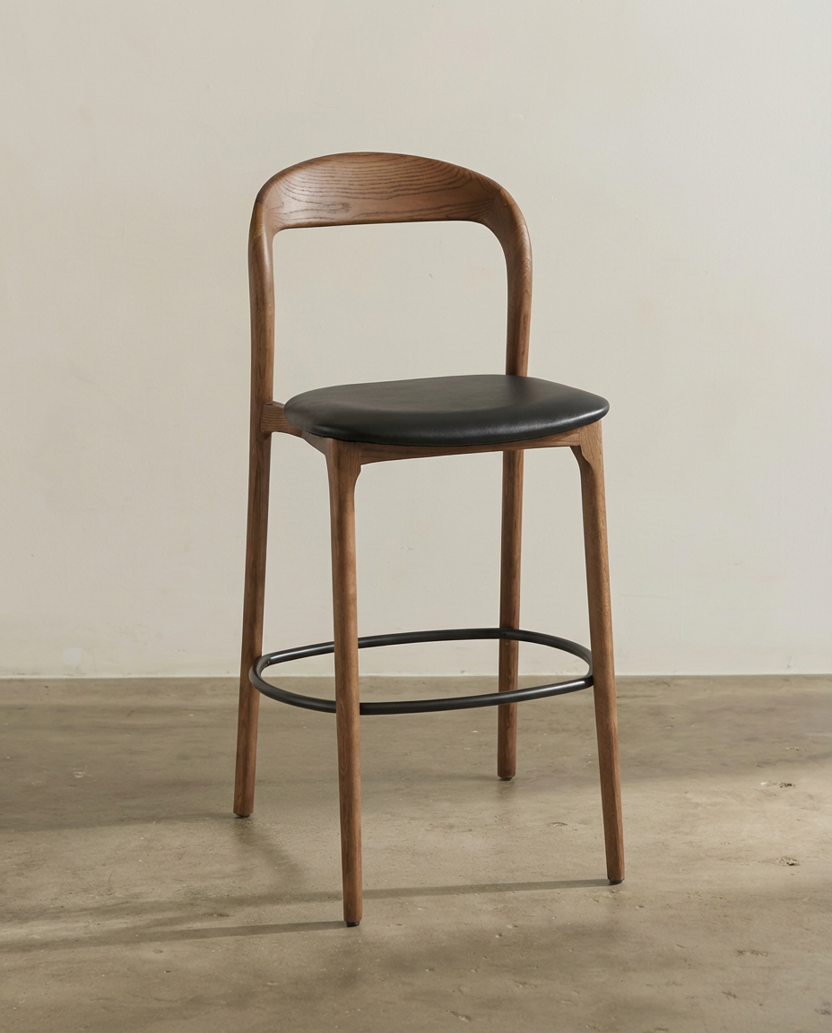 A modern wooden chair with a black cushioned seat and a circular metal footrest, set against a plain wall on a concrete floor.