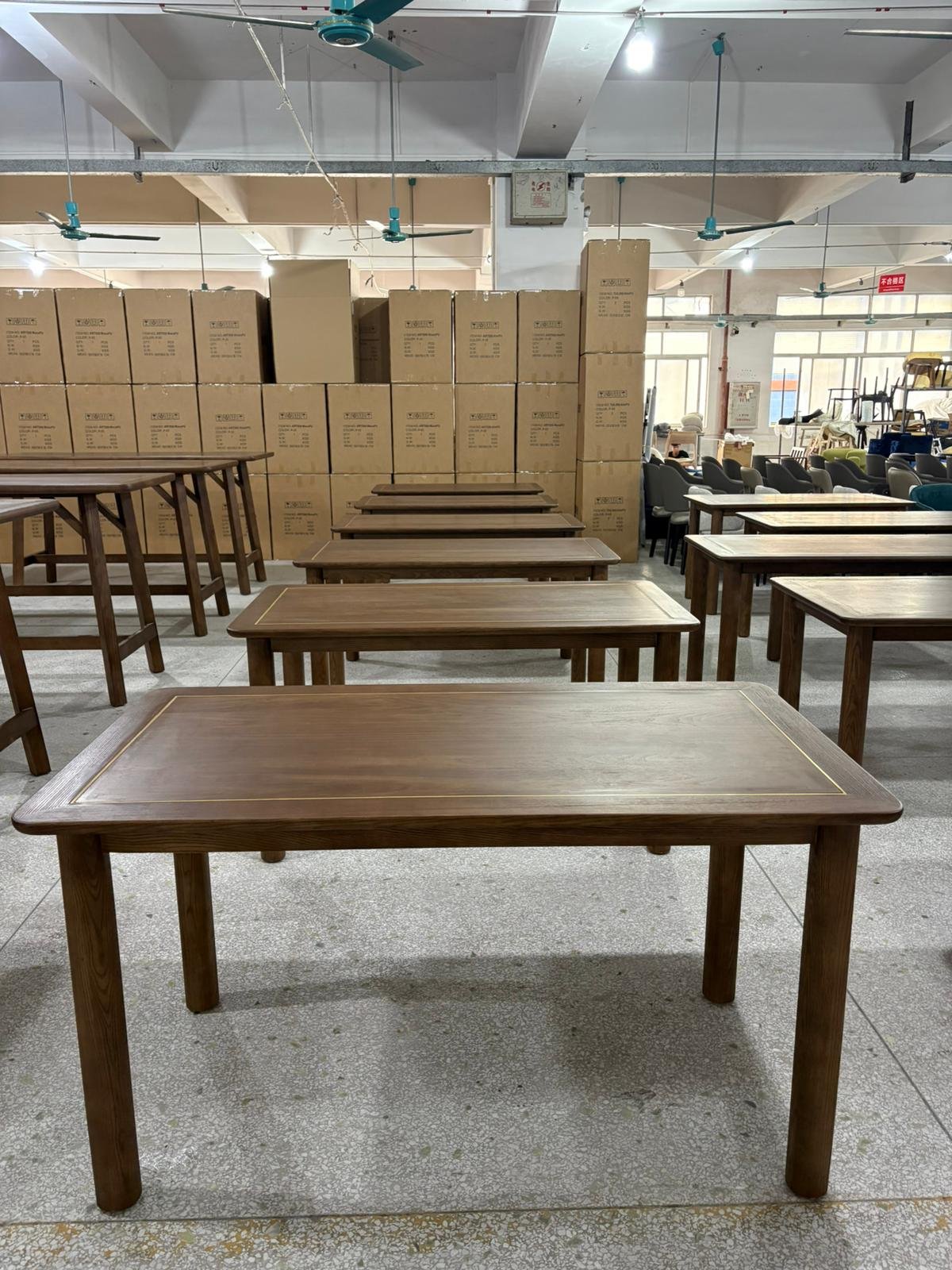 A warehouse or showroom with multiple wooden tables and chairs arranged in rows, stacked cardboard boxes in the background, and ceiling fans overhead.