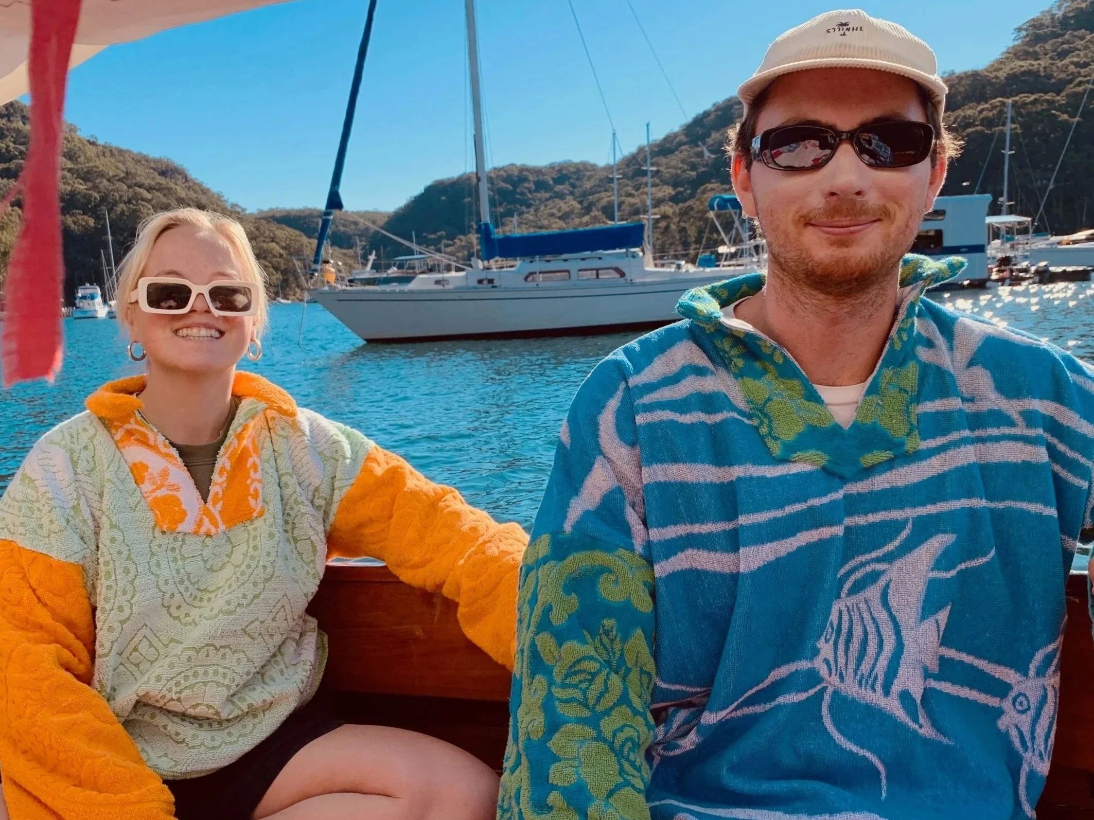 Two people smiling on a boat with boats and hills in the background, wearing colorful warm clothing.