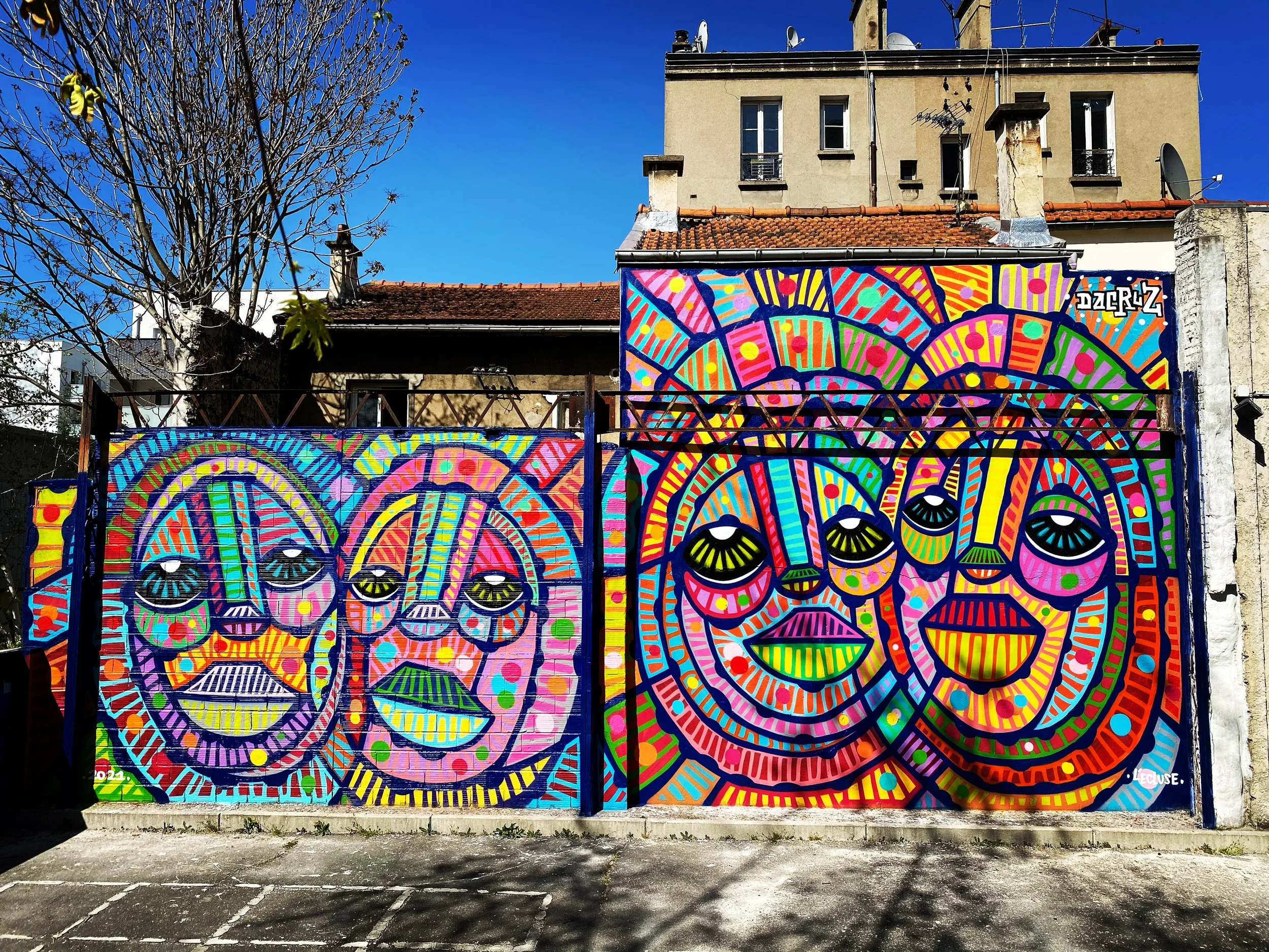 Mosaïque colorée de visages de style abstrait et géométrique sur un mur extérieur, sous un ciel bleu en arrière-plan.