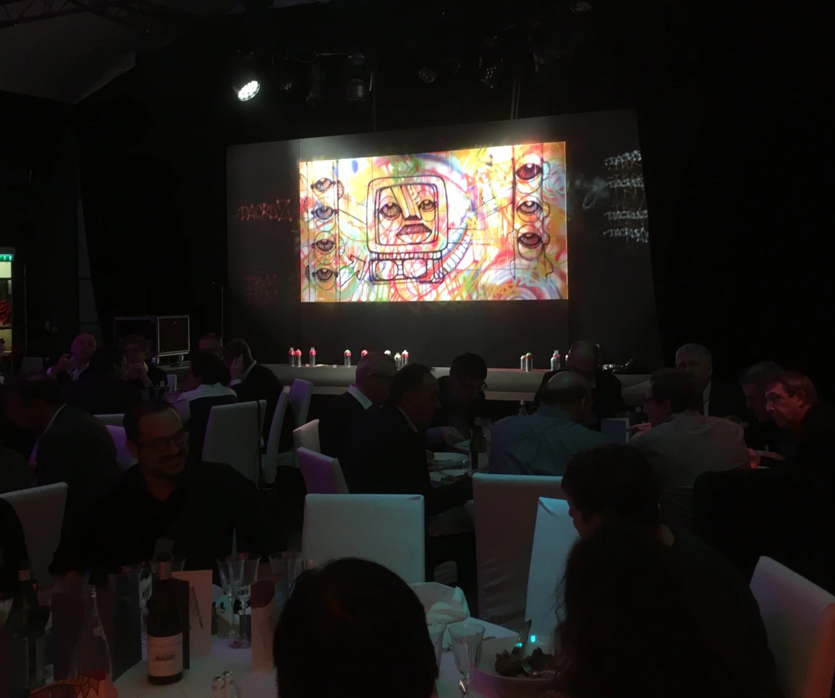 Une scène d'événement avec un grand écran affichant une œuvre d'art colorée représentant un visage avec un casque de plongée, en toile de fond un fond artistique vibrant et des personnes assises à des tables dans une ambiance sombre.