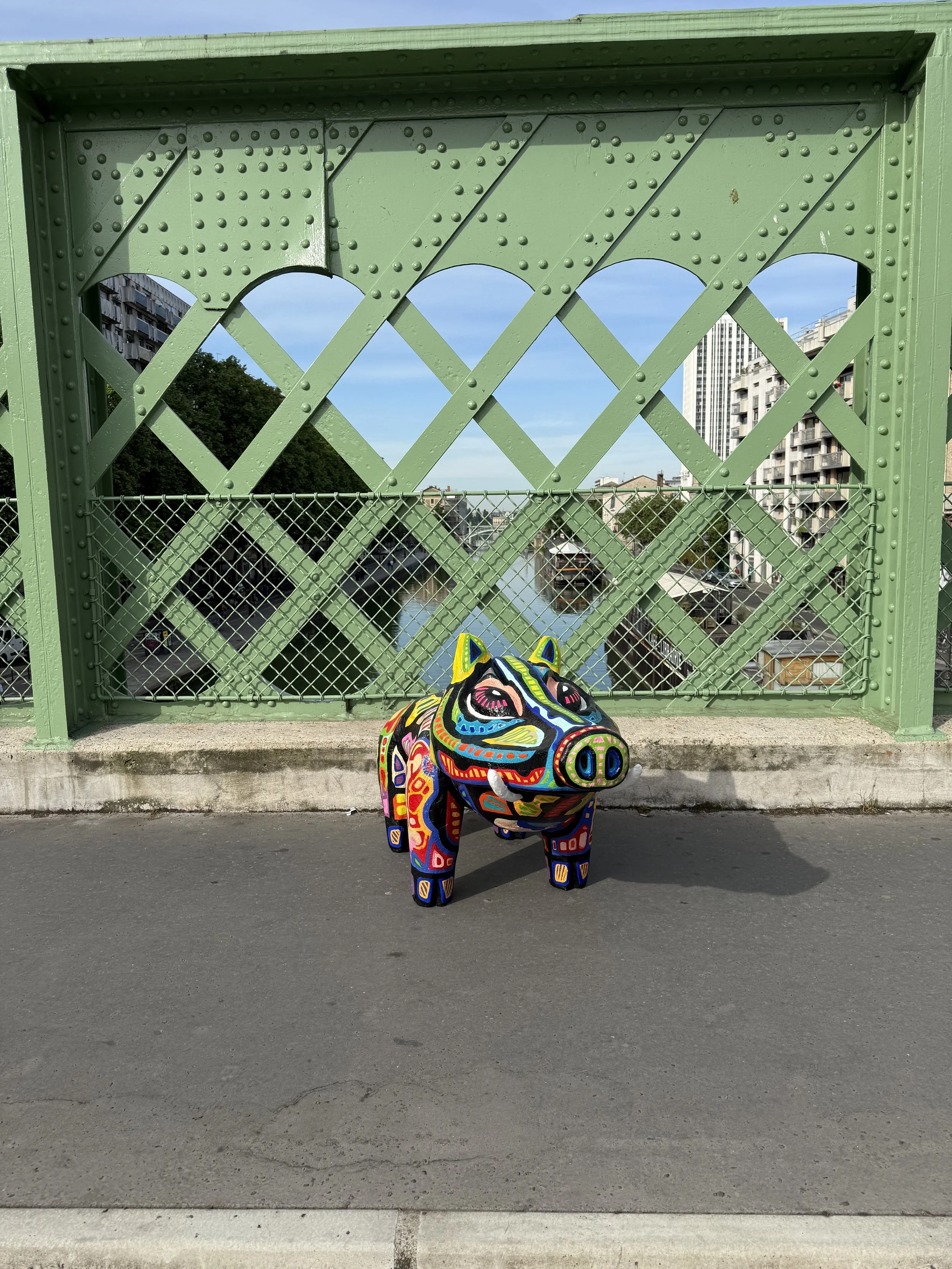 Une sculpture de sanglier coloré et décoratif placé au bord du trottoir avec un pont en fer vert en arrière-plan et un canal avec des bateaux, dans un environnement urbain.