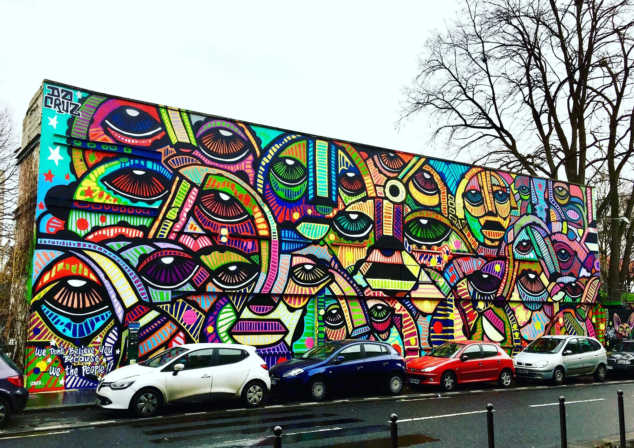 Un mur coloré avec une fresque de style graffiti représentant plusieurs visages stylisés avec des yeux grands et expressifs, accompagnée d'un texte en bas gauche disant "We don't believe you because we the people".