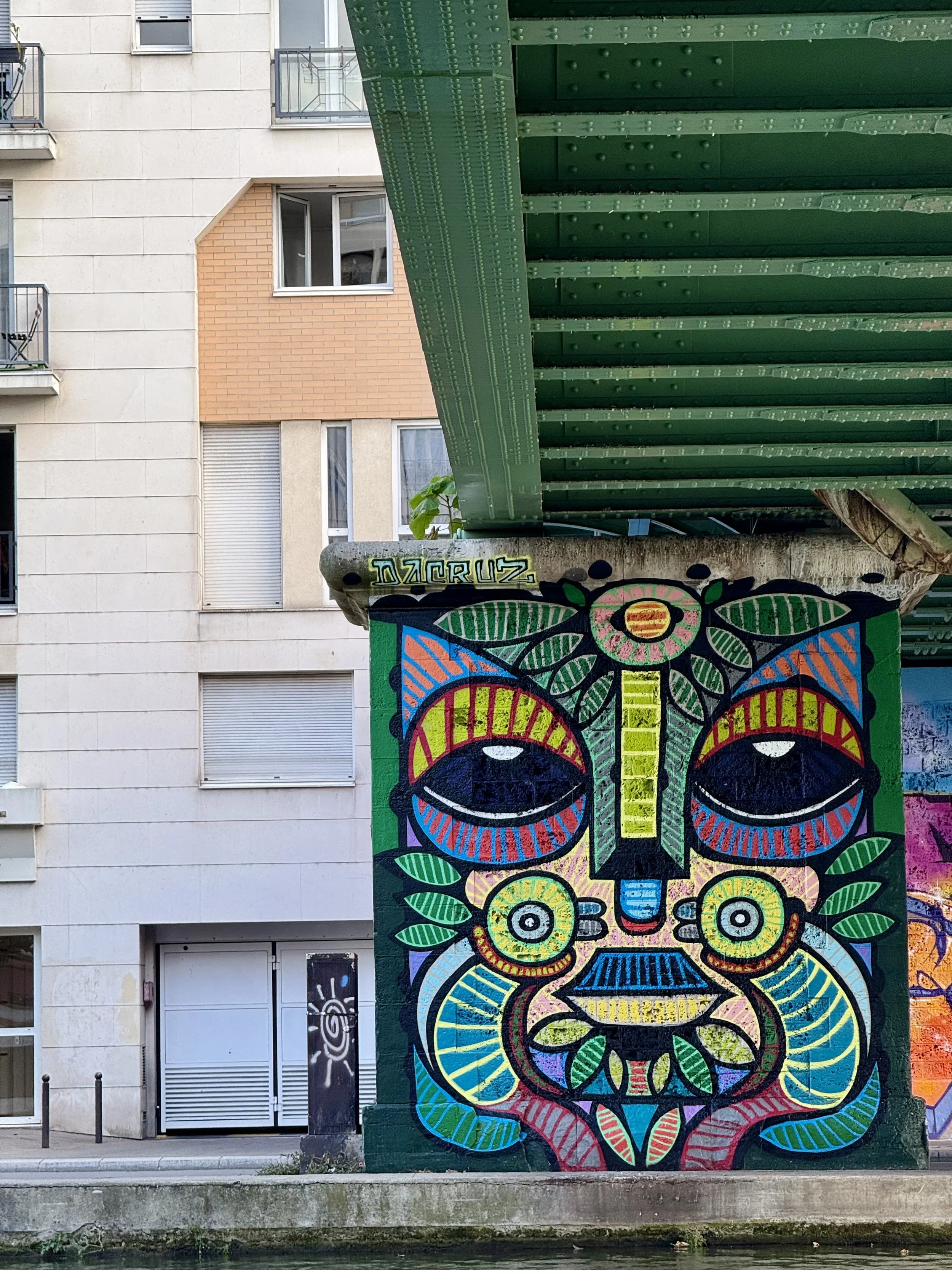 Une fresque de graffiti colorée représentant un visage d'inspiration tribale, avec des motifs symétriques, des yeux grands et expressifs, un nez long et une bouche avec des dents. Il y a aussi des feuilles dessinées autour du visage, le tout peint sur un mur dans un environnement urbain.