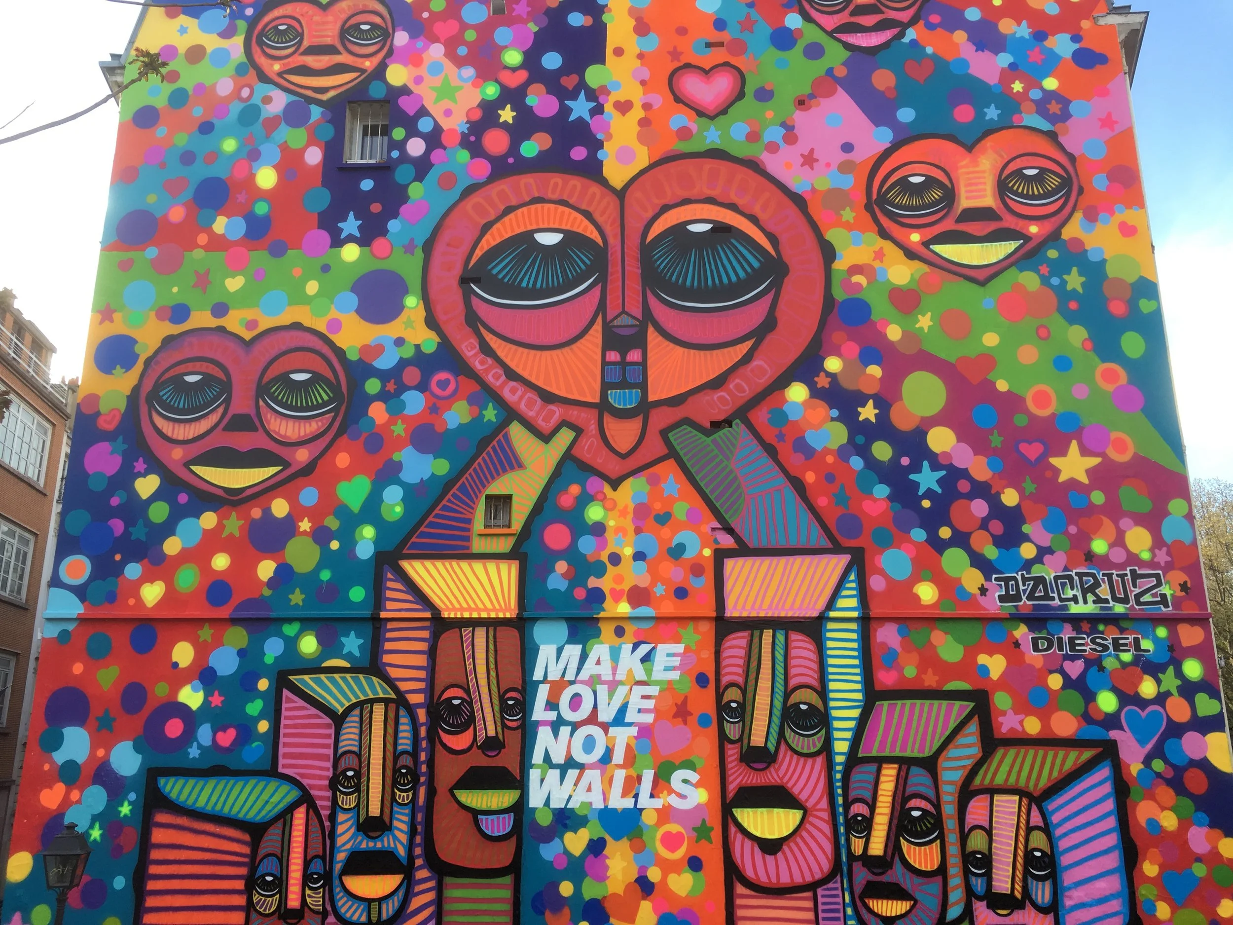 Une fresque murale colorée représentant plusieurs visages stylisés avec de grands yeux, un cœur, des étoiles et des cœurs en fond. La phrase « MAKE LOVE NOT WALLS » est inscrite au centre.
