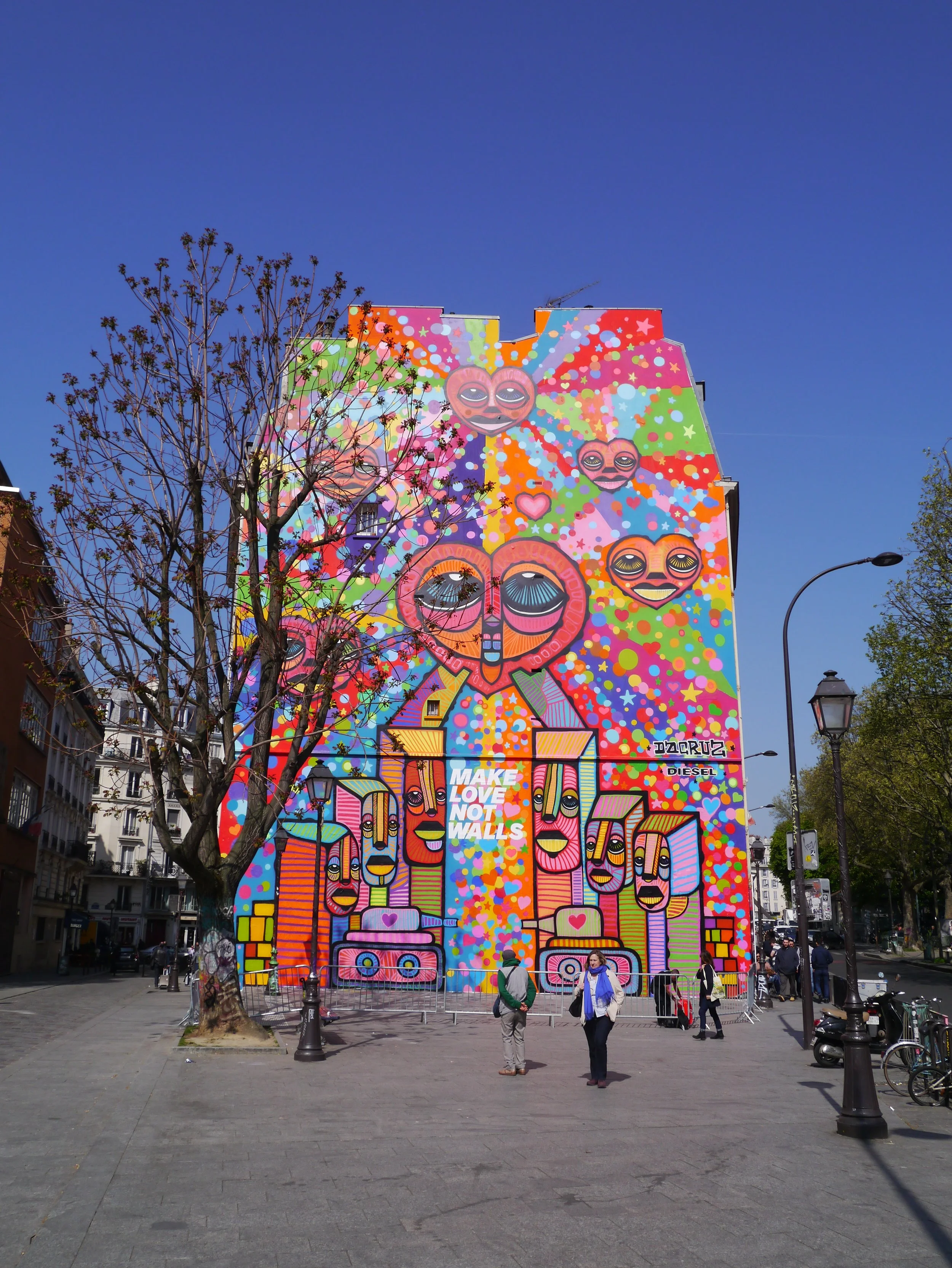 Une grande fresque colorée sur un bâtiment avec des visages souriants, des cœurs, et le texte "MAKE LOVE NOT WALLS".
