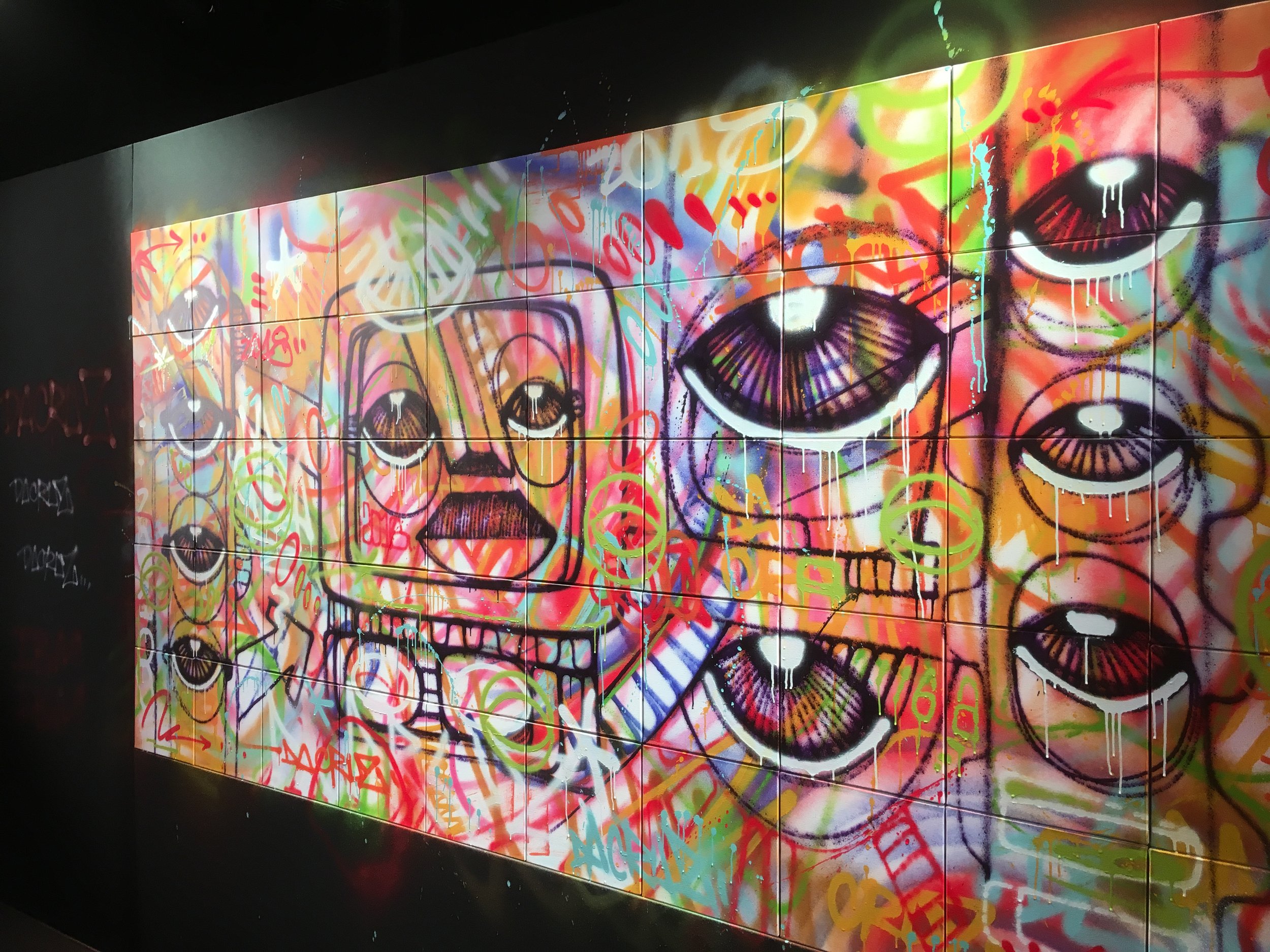 Graffiti coloré de visages stylisés avec des grands yeux et des bouches, intégrant des formes abstraites et des tags. Le tout en couleurs vives comme le vert, orange, violet et rouge.