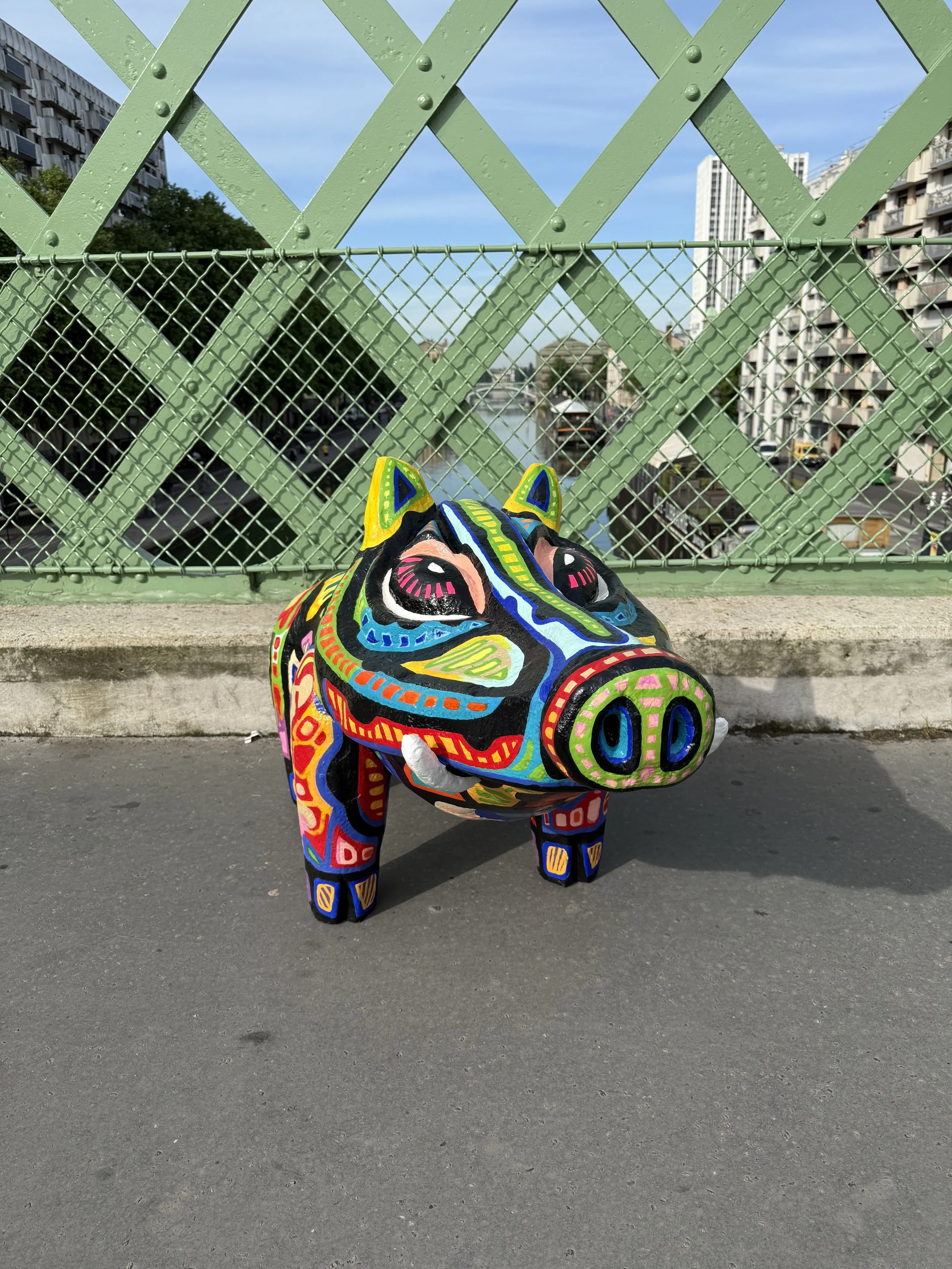 Une sculpture de sanglier colorée et artistique posée sur le trottoir, avec un pont vert et une ville en arrière-plan.