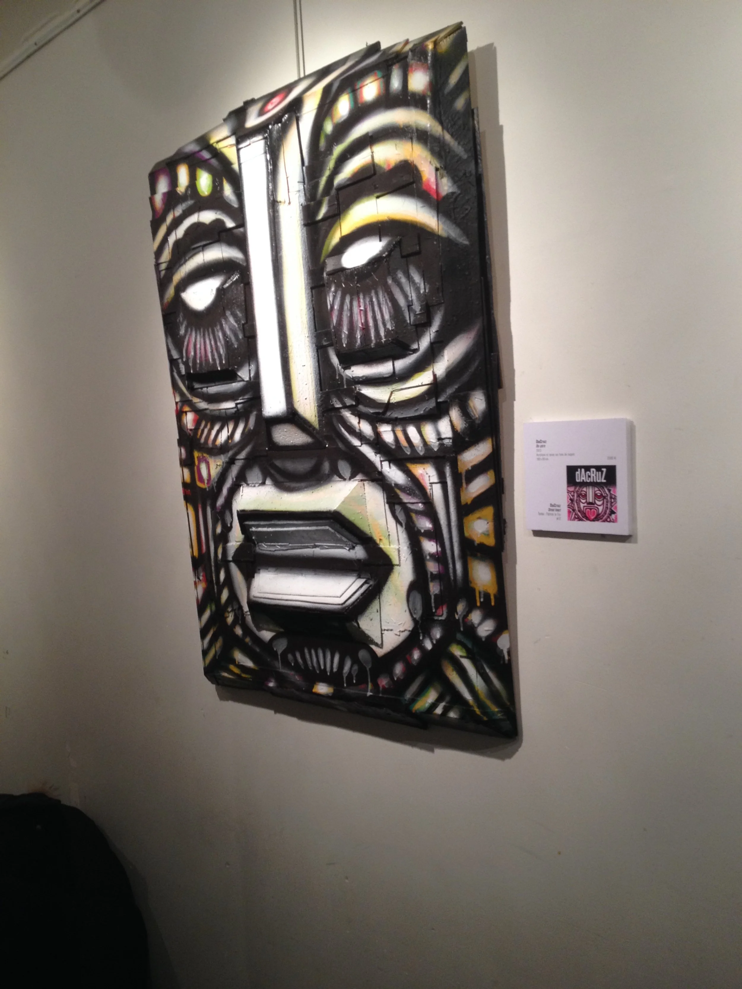 Une peinture murale de style graffiti représentant un visage humain stylisé, avec de grands yeux, un nez long et des lèvres closes, sur une toile. La peinture utilise des couleurs sombres avec des accents de couleurs vives.