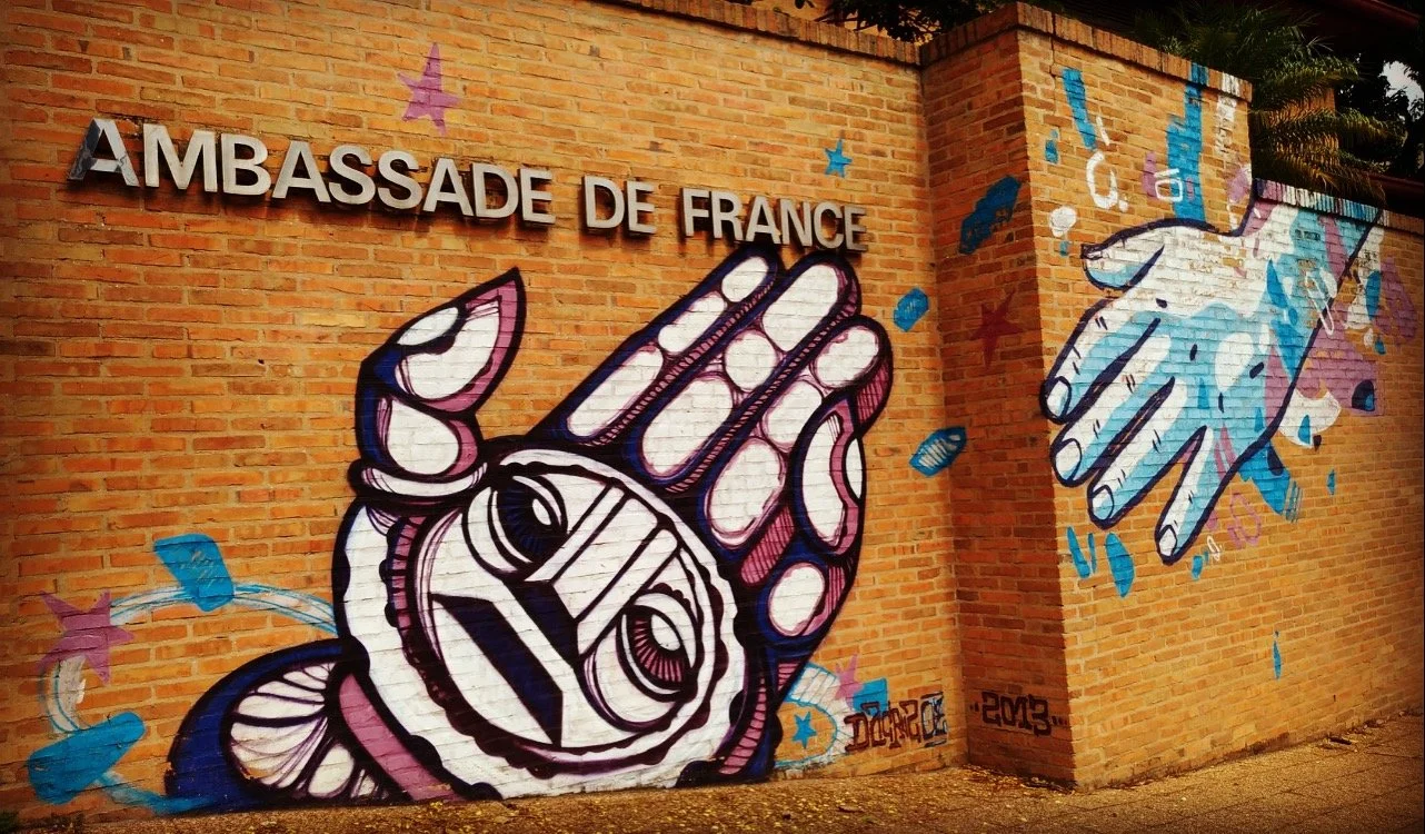 Une fresque murale colorée représentant une main avec des étoiles et un éclair, à côté de la mention "Ambassade de France" sur un mur en briques, avec un poisson et une main étoilés.