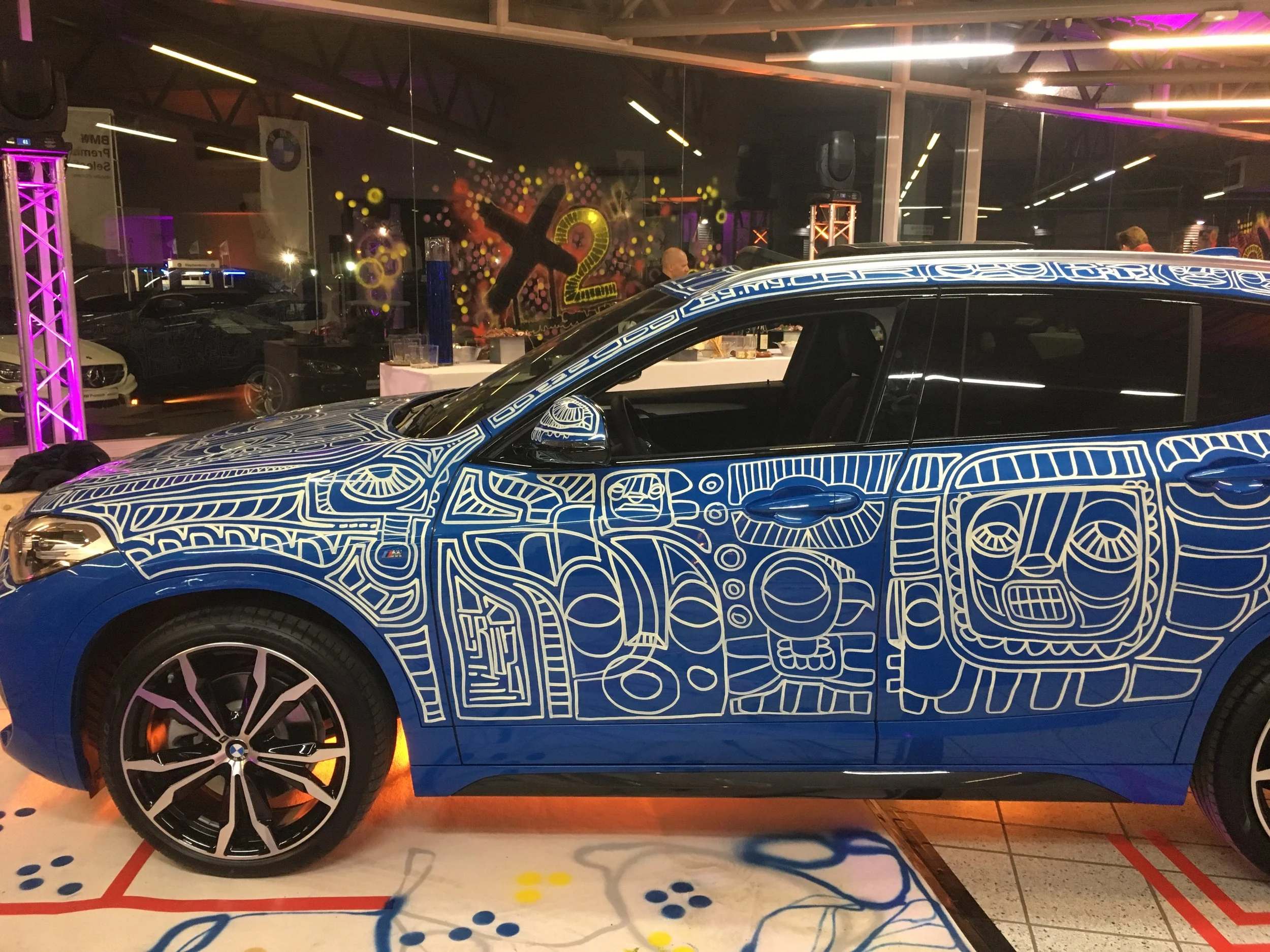 Voiture électrique de couleur bleue avec un design artistique en lignes blanches représentant des visages et des motifs abstraits, exposée lors d'un événement à l'intérieur d'une salle.