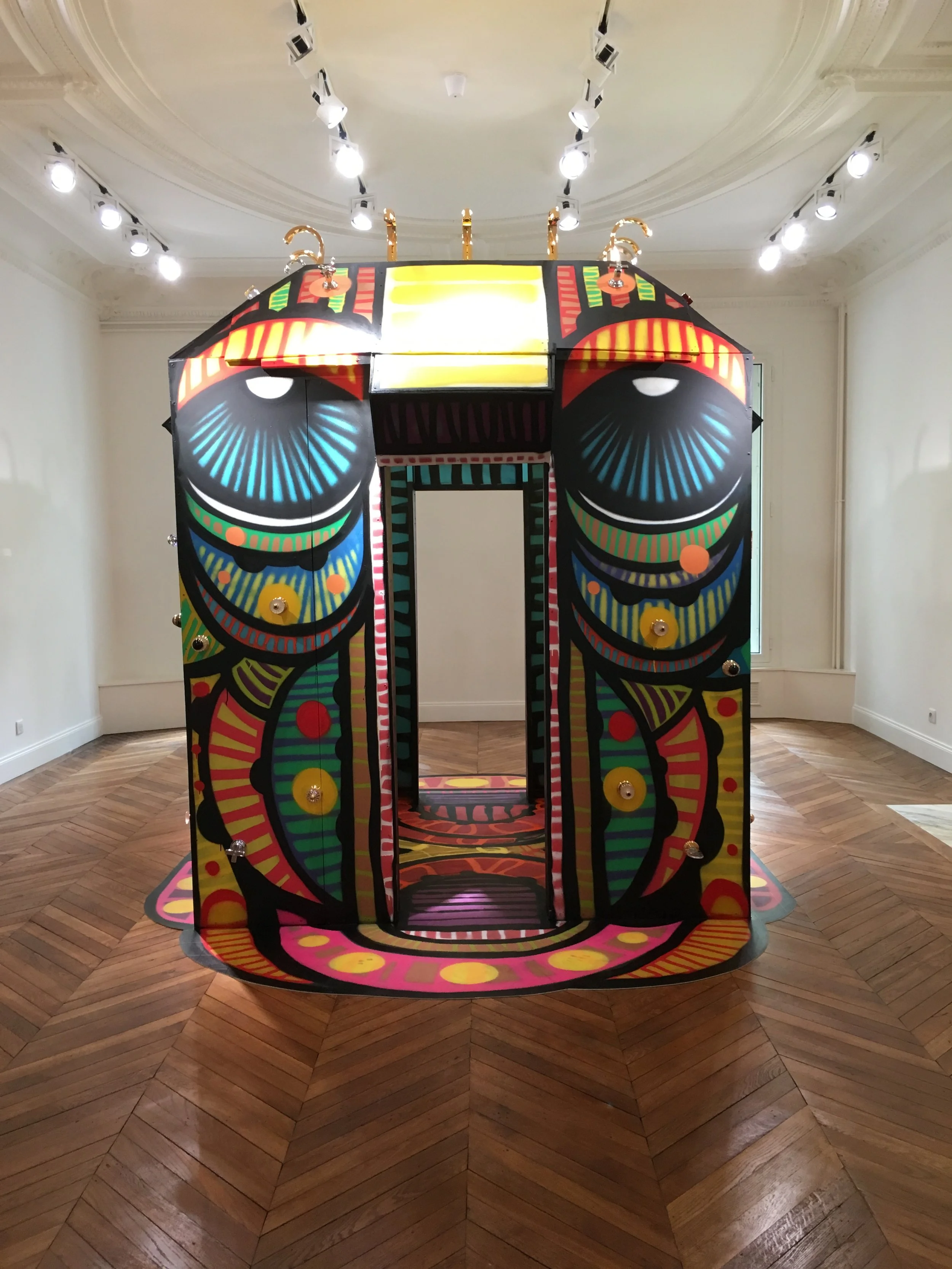 Une installation artistique de forme ronde et colorée avec des motifs graphiques, située dans une pièce à parquet en bois et éclairée par des lumières au plafond.