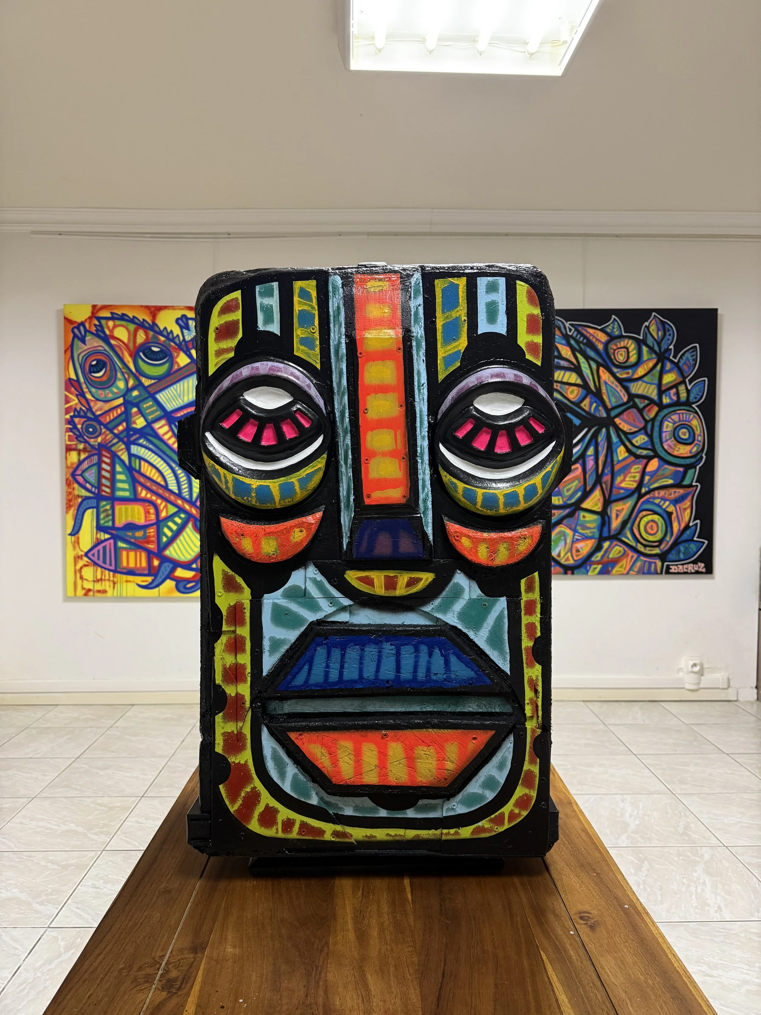 Une sculpture ou panneau en forme de visage stylisé, peint avec des couleurs vives et des motifs abstraits, placée sur une table en bois, avec deux peintures colorées en arrière-plan dans un espace d'exposition.