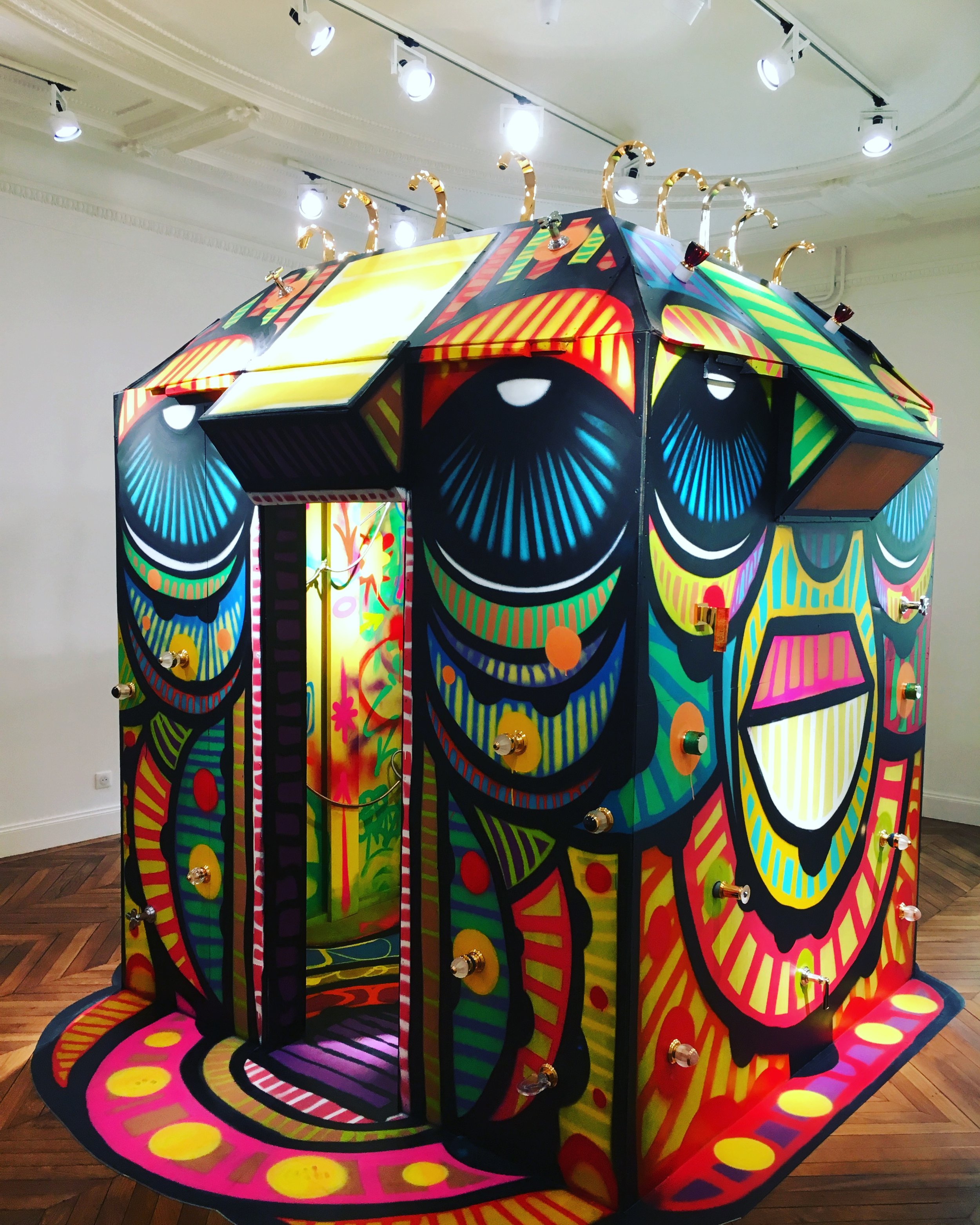 Une petite maison en bois colorée avec un motif artistique et des lumières suspendues au plafond, située dans une galerie d'art ou un musée.