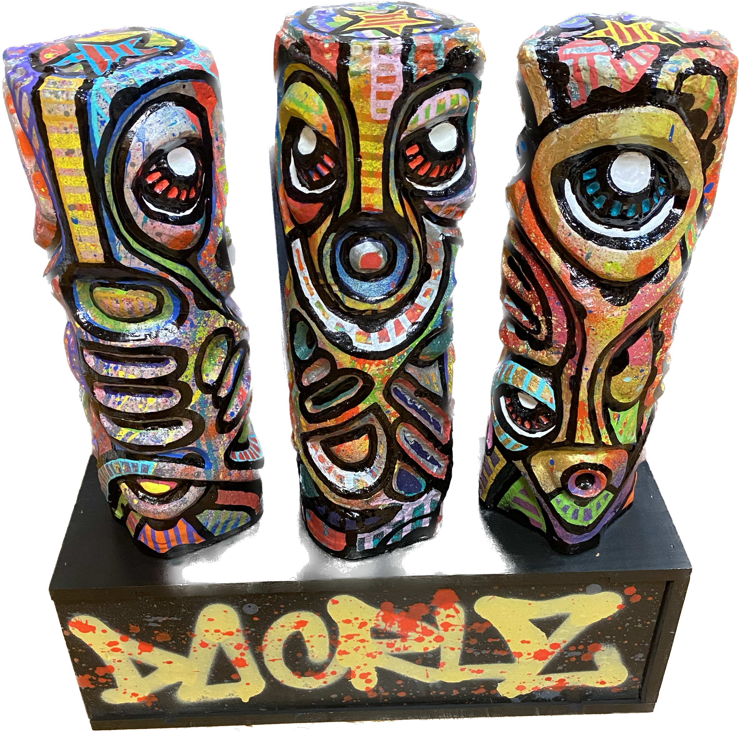 Trois sculptures de style tribal ou indigène, peintes de couleurs vives, représentant des visages ou des masques, dressées sur une plateforme noire avec le mot 'ZANKA' en graffiti jaune et rouge.