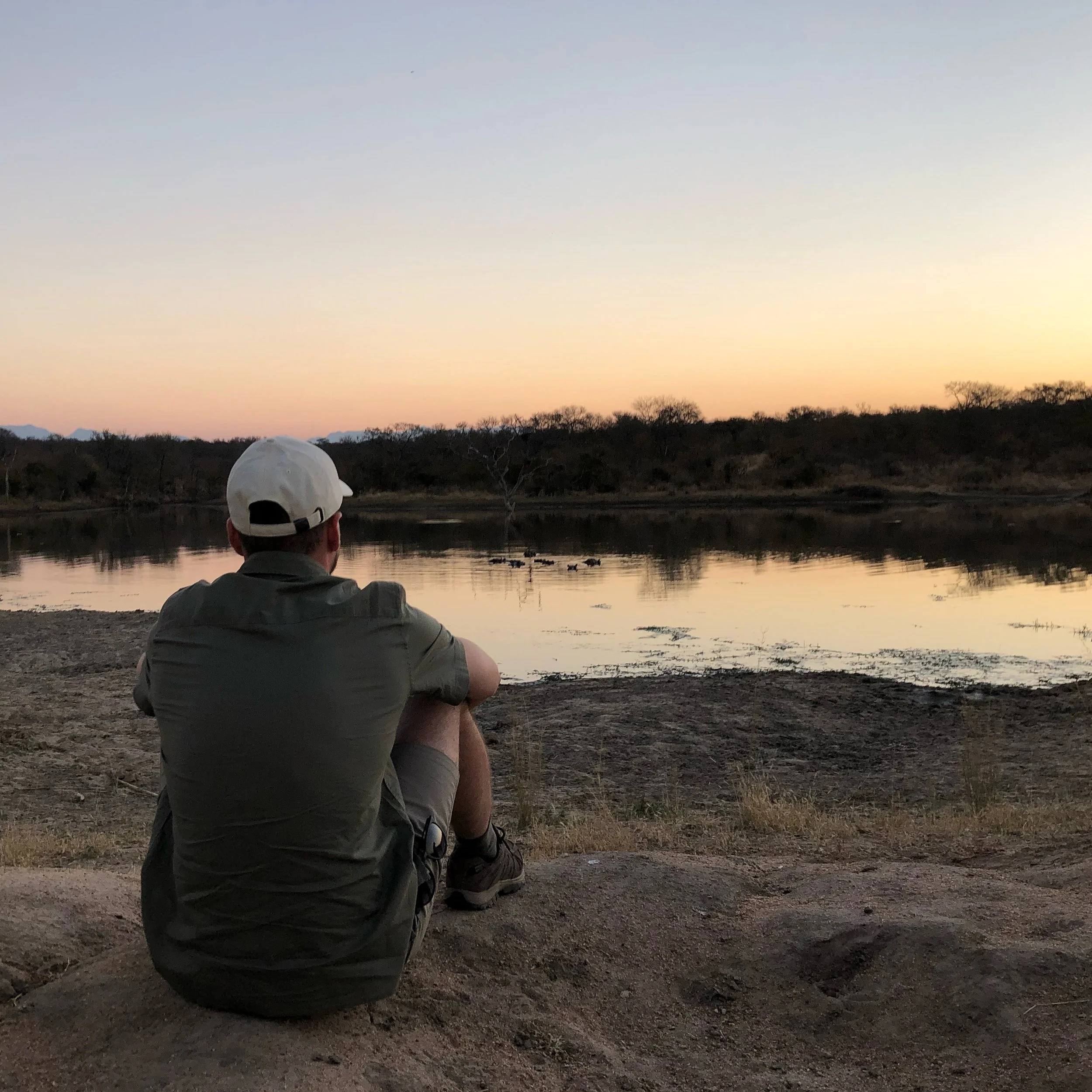 Un homme assis sur la rive d'un lac, regardant le coucher de soleil, avec des arbres et des montagnes à l'horizon.