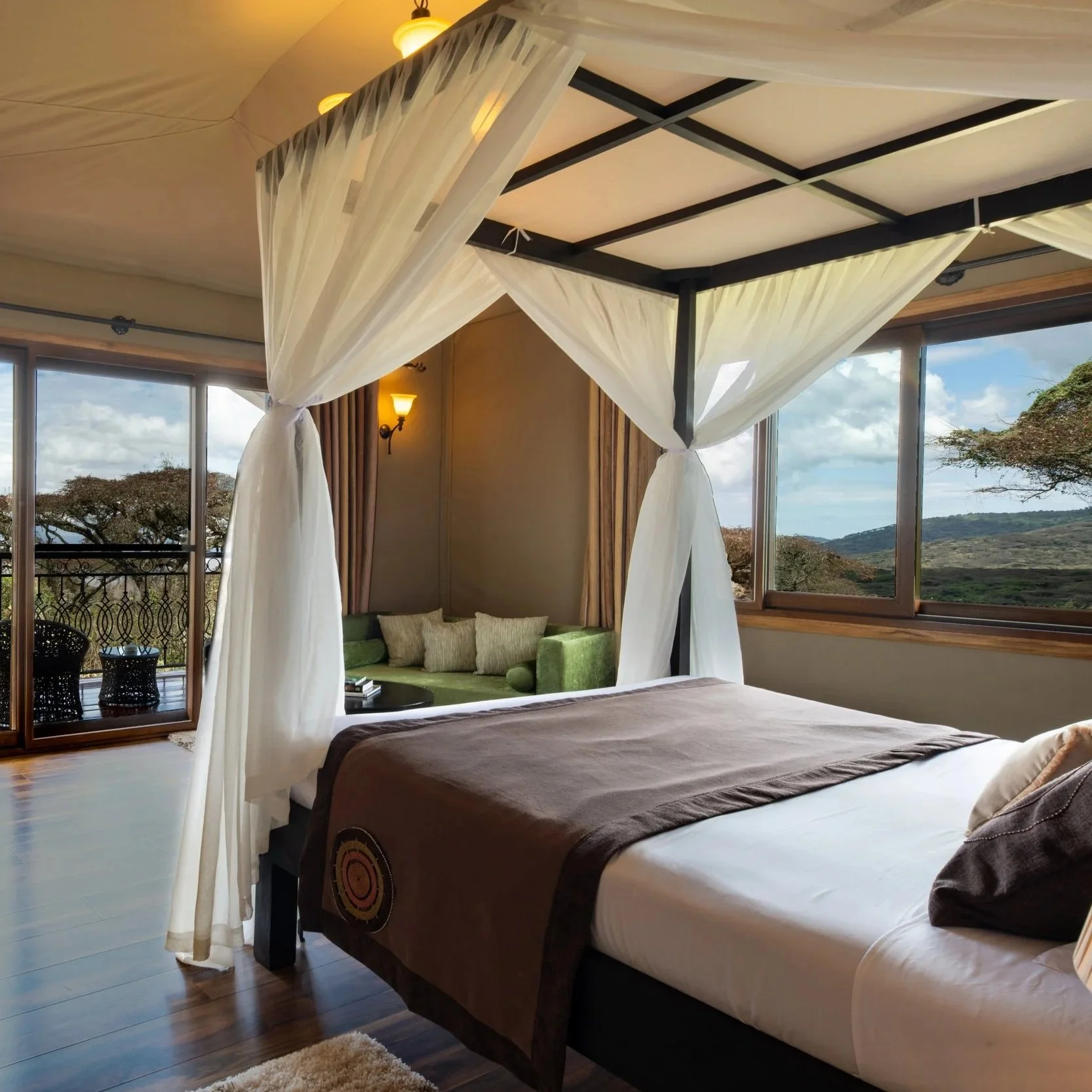 Chambre avec lit à baldaquin, vue sur un paysage naturel avec arbres et montagnes, fenêtre large, coin salon avec canapé vert et coussins, décor chaleureux avec lampes et rideaux.