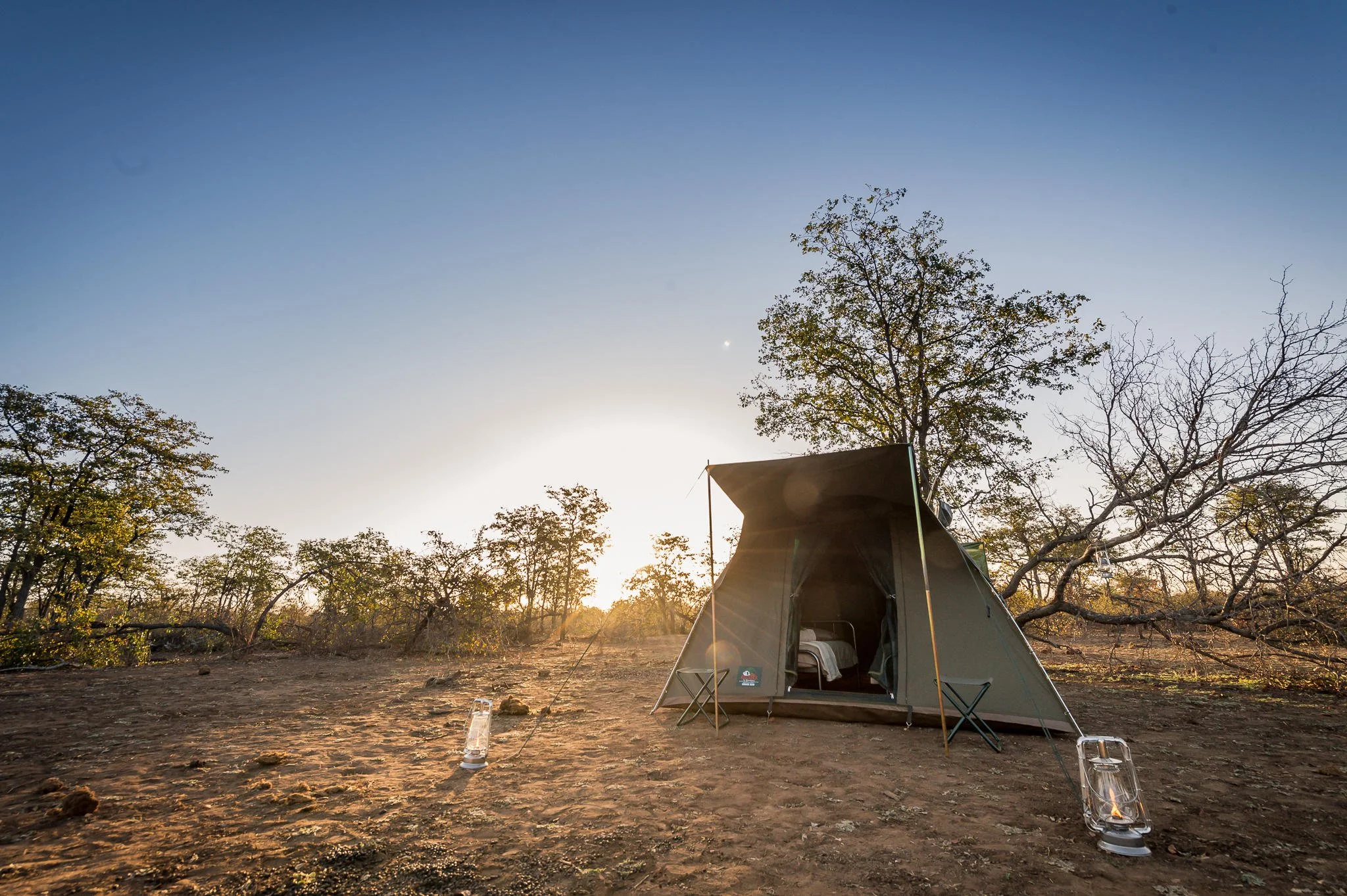 Une tente de camping dans un paysage désertique au coucher du soleil, entourée de quelques arbres et lampes à pétrole au sol.