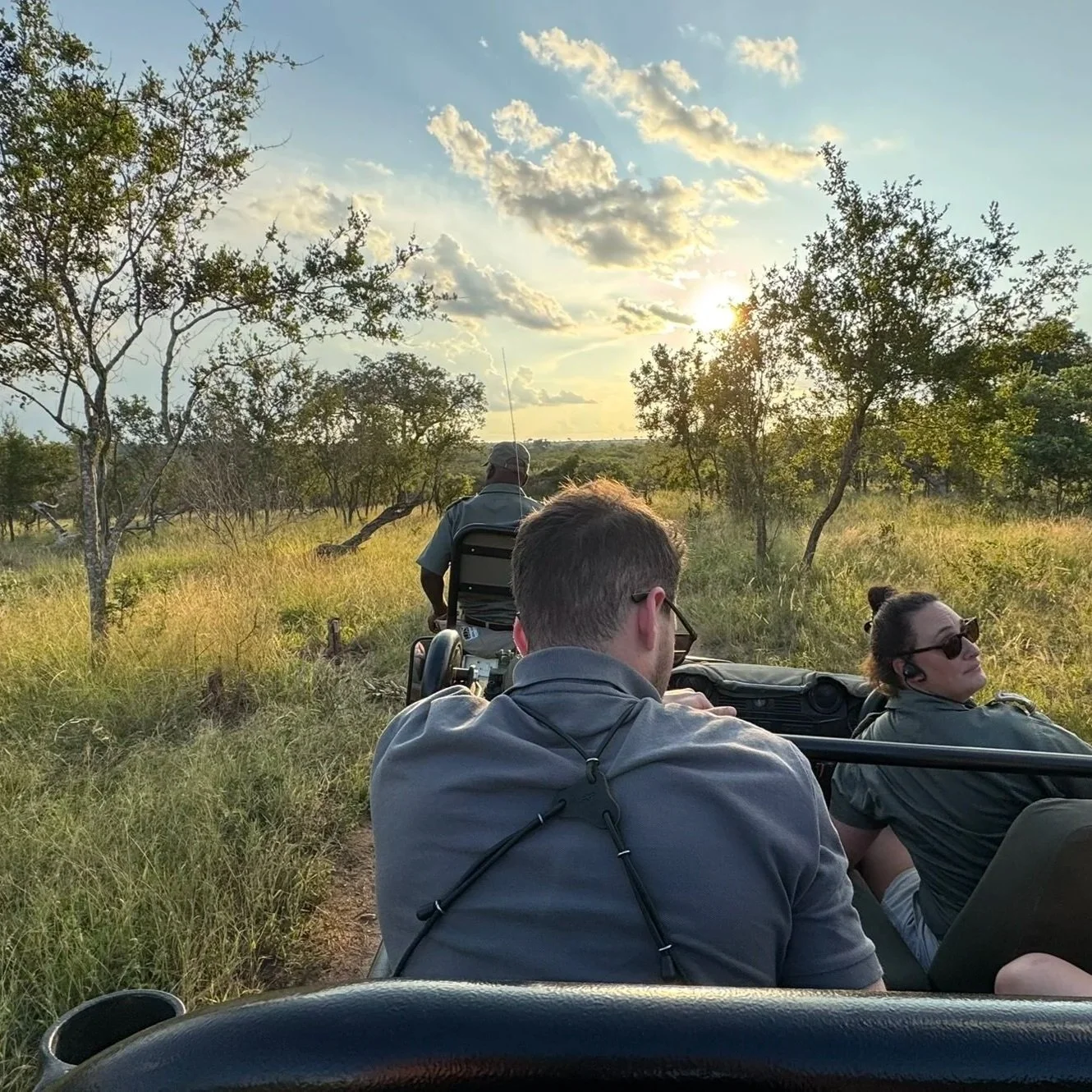 Trois personnes faisant un safari en véhicule à ciel ouvert dans une savane, avec des arbres et un ciel partiellement nuageux au coucher du soleil.