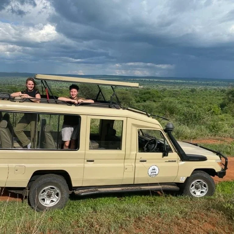 Deux personnes dans un véhicule de safari, un homme et une femme, près de la nature avec un ciel nuageux en arrière-plan.