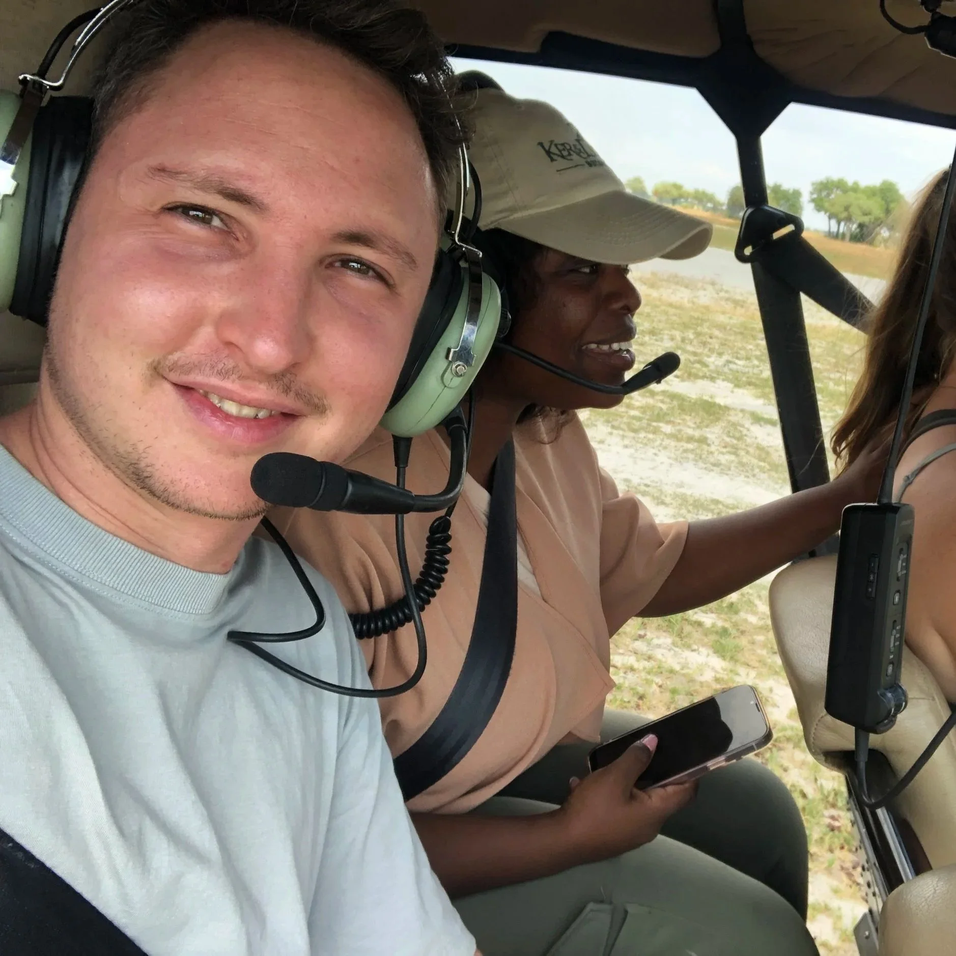 Deux personnes dans un hélicoptère, une jeune homme souriant et une femme avec un casque de communication, en plein vol au-dessus d'un paysage de nature.