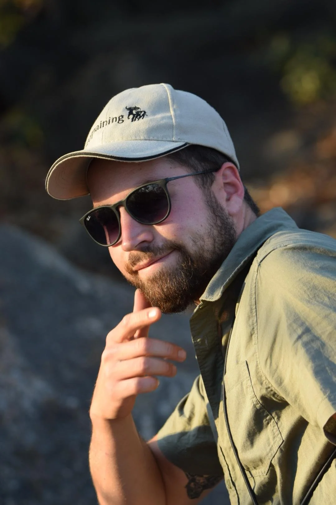 Un homme avec une barbe, portant des lunettes de soleil, un casquette beige et une chemise verte, posant en extérieur.