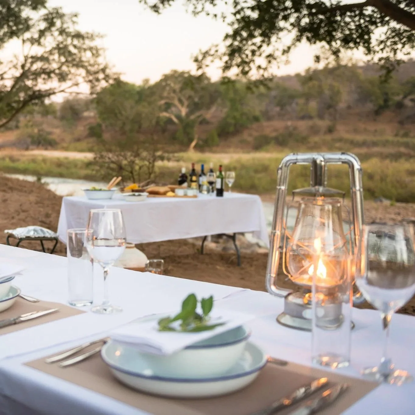 Table dressée pour un dîner en plein air avec lanternes, assiettes, verres, et bouteilles de vin au coucher du soleil dans un environnement naturel avec des arbres et un ruisseau.
