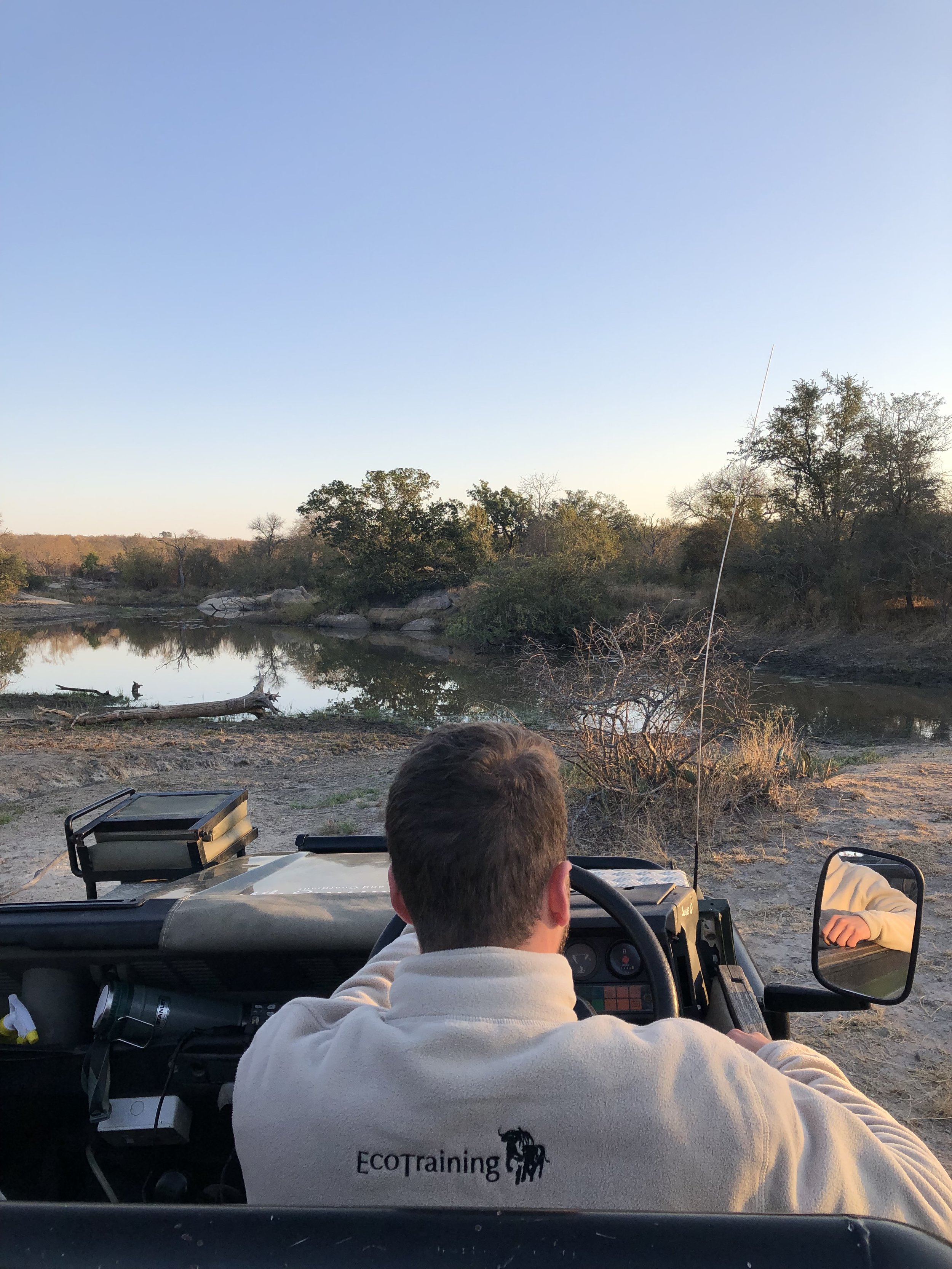 Une personne vue de dos, assise dans une jeep, regarde un paysage de rivière et de nature avec des arbres et un ciel clair, en fin d'après-midi.