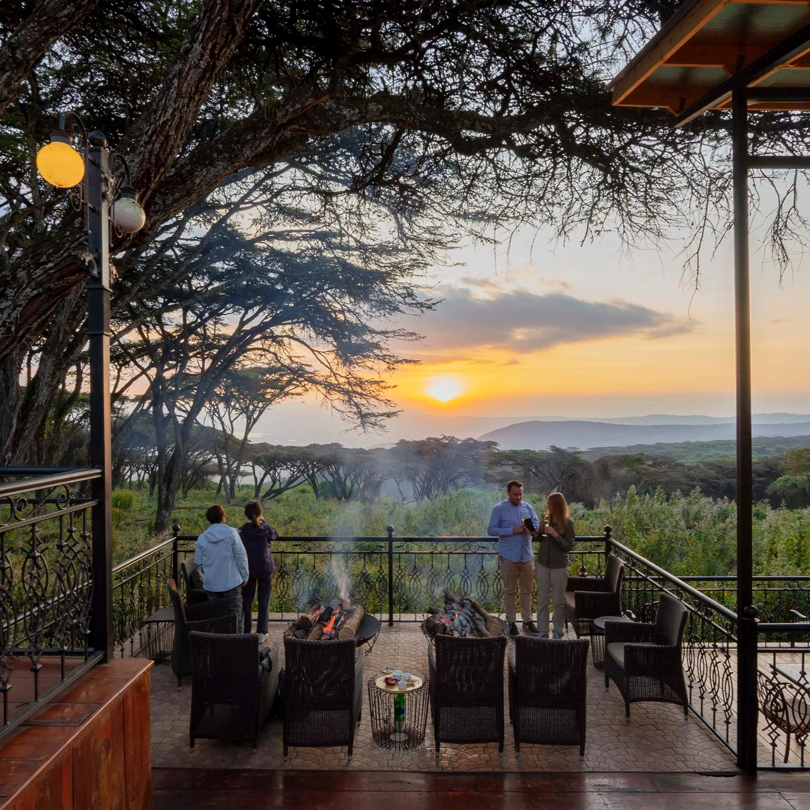 Groupe de personnes appréciant un coucher de soleil sur une terrasse entourée de nature, avec du mobilier en bambou et un feu de camp.