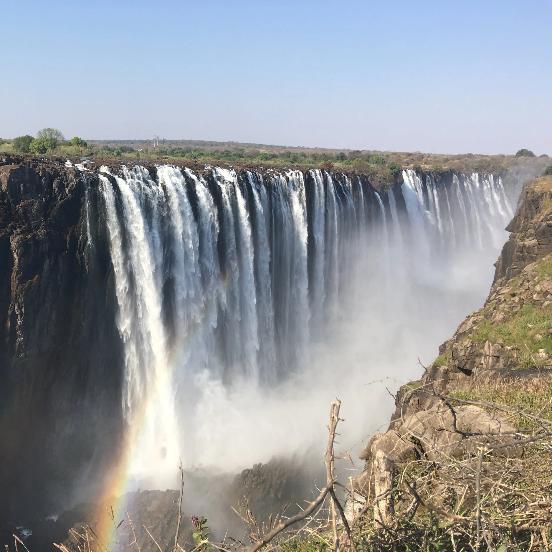 Chute d'eau massive avec un arc-en-ciel dans la brume, vue depuis le bord du précipice, en plein jour avec un ciel bleu clair.