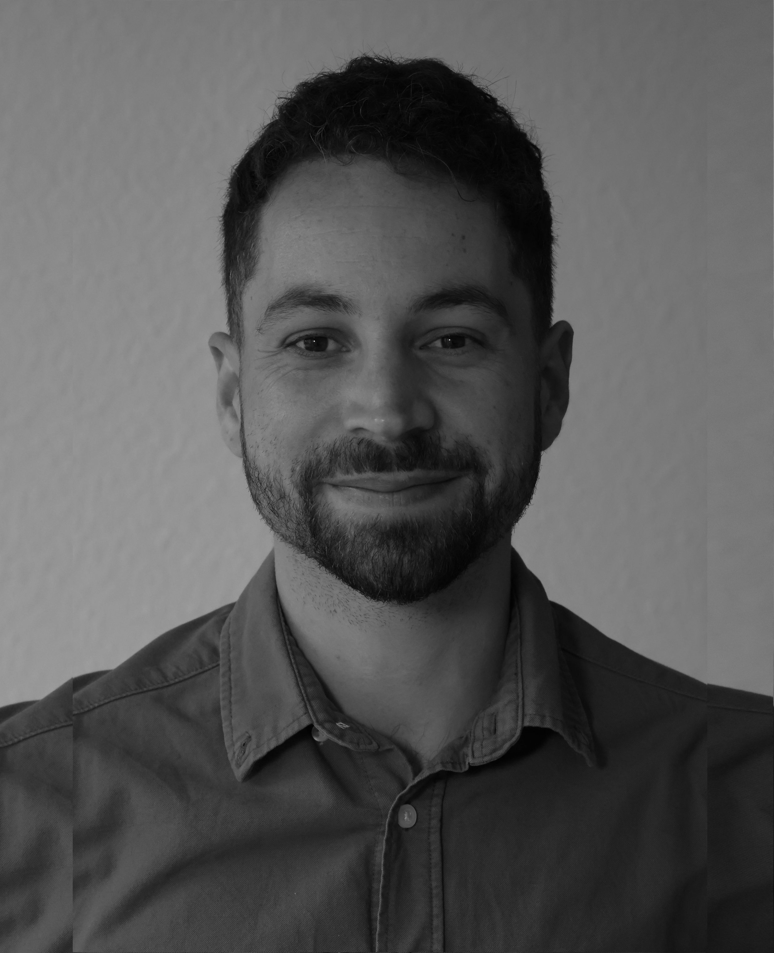 Portrait en noir et blanc d'un homme souriant, portant une chemise à col boutonnière, avec un fond neutre.