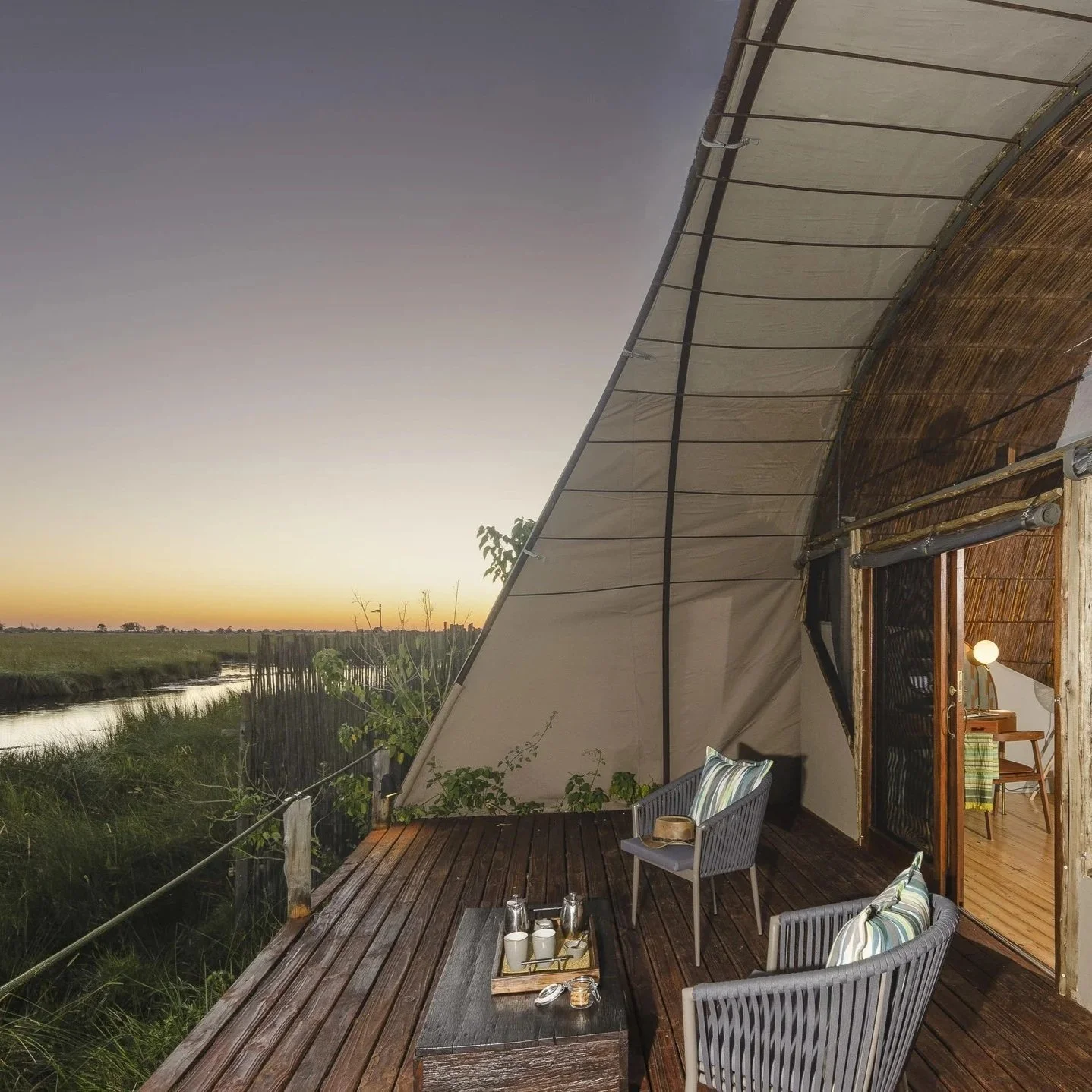 Terrasse en bois avec deux fauteuils en rotin, coussins, table avec boissons, et vue sur un coucher de soleil sur un paysage naturel avec une rivière.