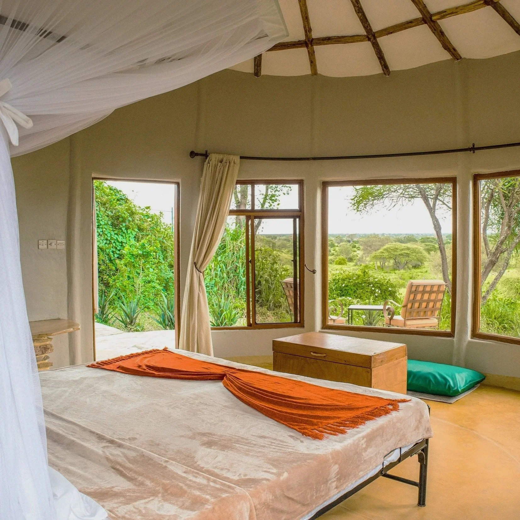 Chambre avec grandes fenêtres offrant une vue sur la nature, lit avec drap beige et écharpe orange, coffre en bois, coussin vert, fauteuil en extérieur visible par la fenêtre.