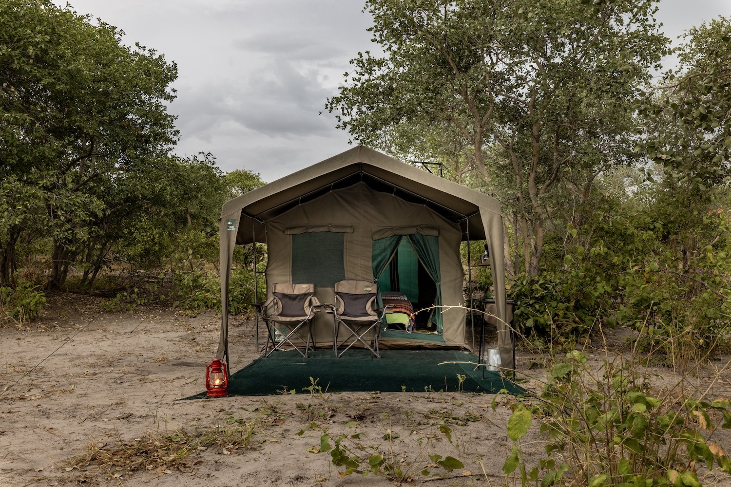 Tente de camping avec deux chaises, lampe à pétrole rouge, et matériel de camping sur une plateforme dans un environnement naturel avec des arbres et un ciel nuageux.