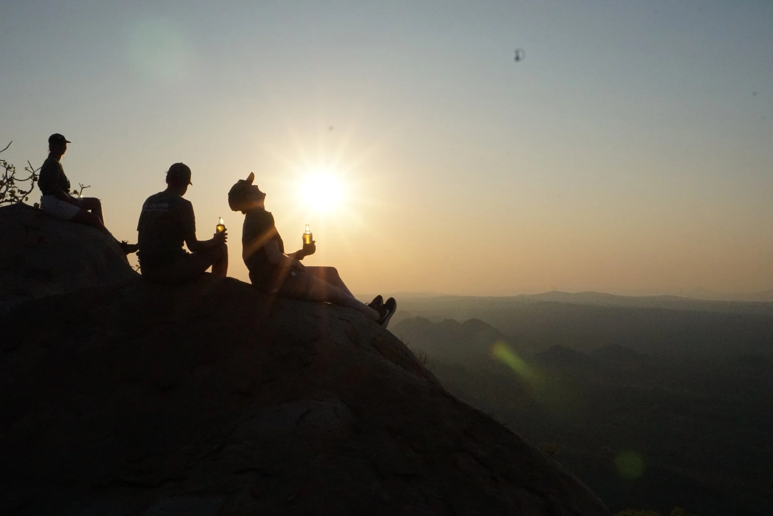 Silhouettes of four people sitting on a rock at sunset, with one person holding a bottle.